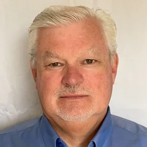 A man wearing a blue collared shirt, looking directly at the camera against a light background.
