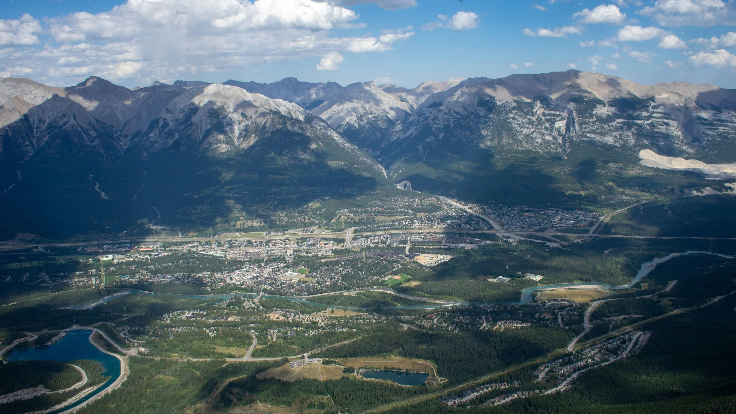 Aerial view of a mountain range with snow-capped peaks, a valley with a town, highways, and a river, under a partly cloudy sky in Colorado.