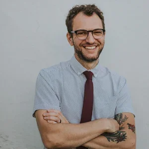 A man with glasses, a beard, and curly hair smiling with arms crossed, wearing a light blue shirt, a dark red tie, and showing tattoos on his right forearm, standing against a plain white wall.