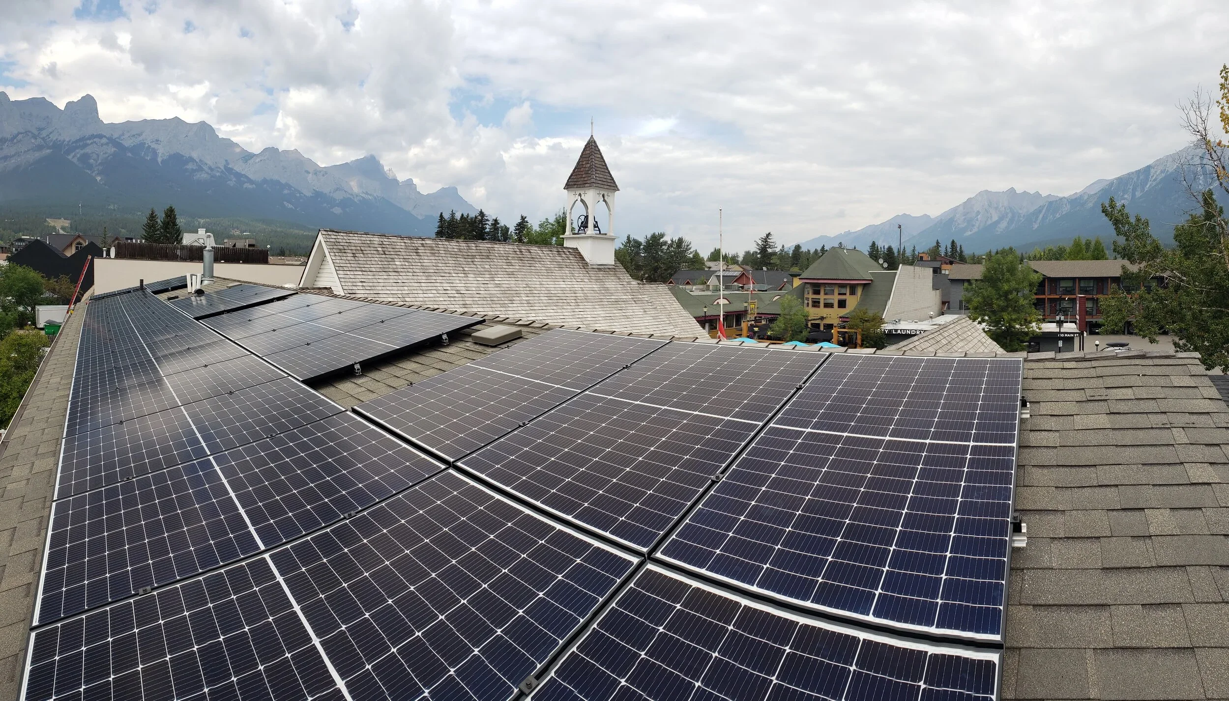 Roof with solar panels overlooking a town with a church, trees, and mountains in the background under a cloudy sky.