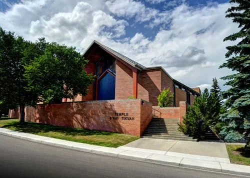 Exterior of a modern brick synagogue with stairs, trees, and a cloudy sky.