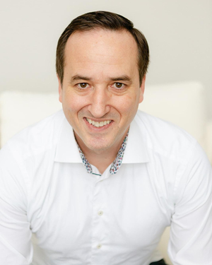 Close-up of a smiling man in a white shirt against a neutral background.