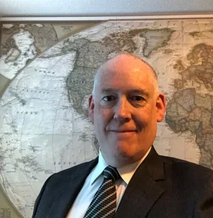 A man with a shaved head wearing a suit and tie, standing in front of a world map.