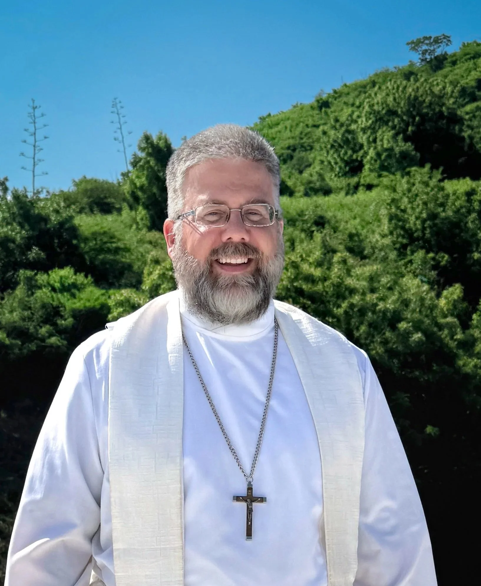 Un pastor luterano con barba y gafas religiosas, viste un hábito blanco y lleva un crucifijo de collar, de fondo hay árboles y un cielo azul.