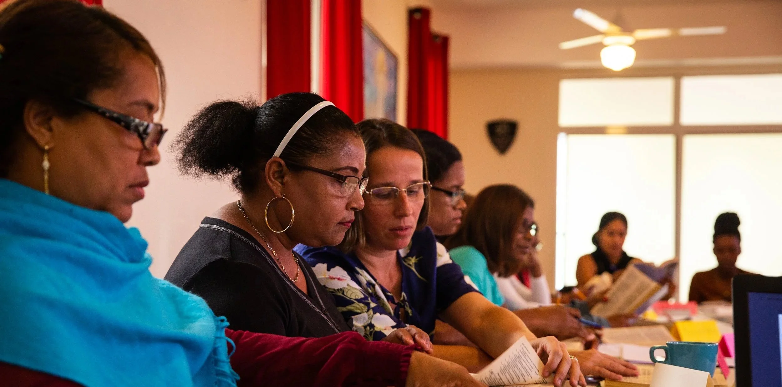 Grupo de mujeres sentadas en una mesa, leyendo documentos en una sala con ventanas grandes y cortinas rojas.