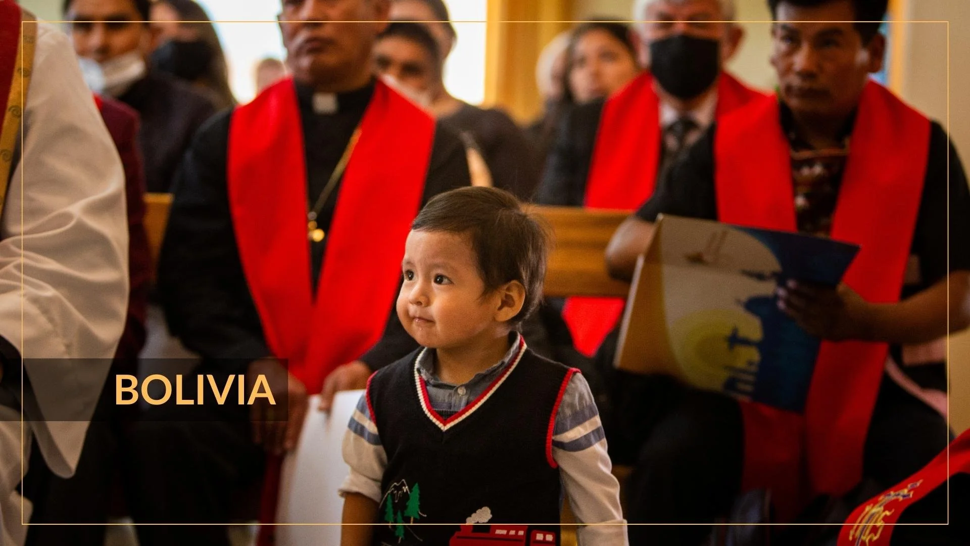 Niño pequeño con barba de triángulo en el pecho, de pie en una ceremonia religiosa con personas adultas vestidas con ropa formal y bufandas rojas, en Bolivia.
