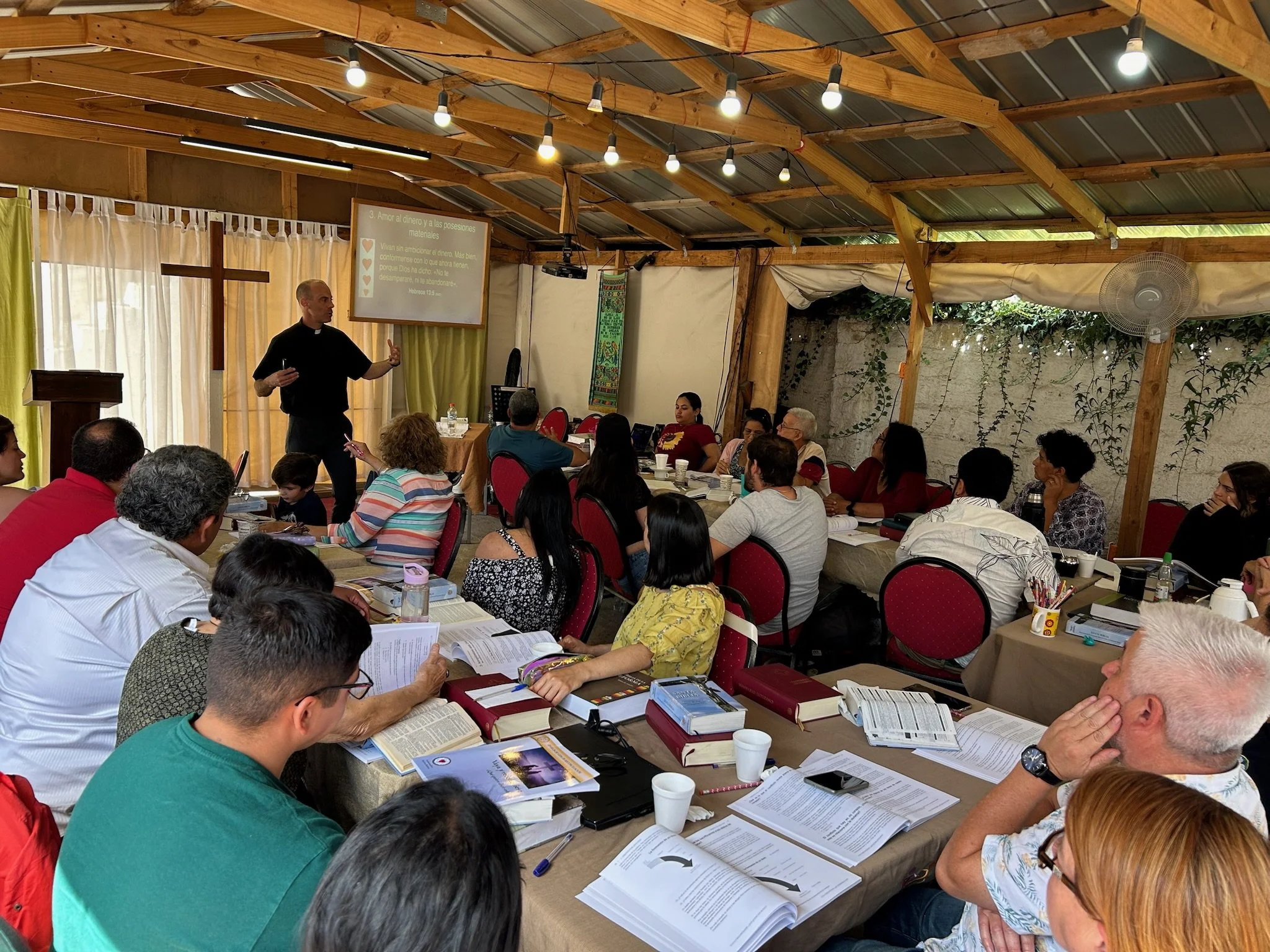 Un pastor dando una charla a un grupo de personas en un recinto techado con madera y luz artificial, con una cruz en la cortina y una pantalla de proyector