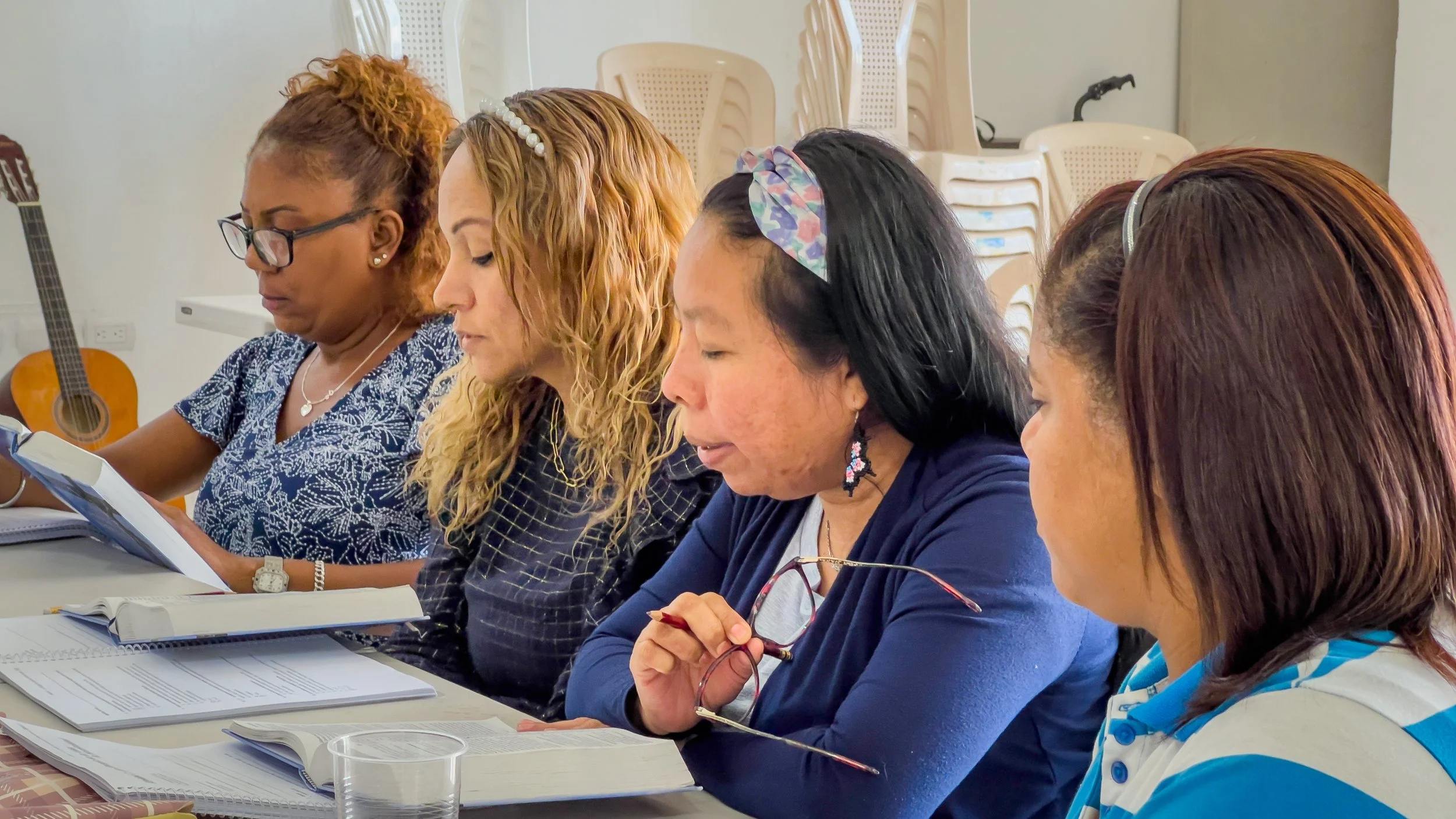 Cinco mujeres sentadas en una mesa, leyendo y tomando notas, en un entorno de reunión o clase, con documentos y libros delante de ellas.