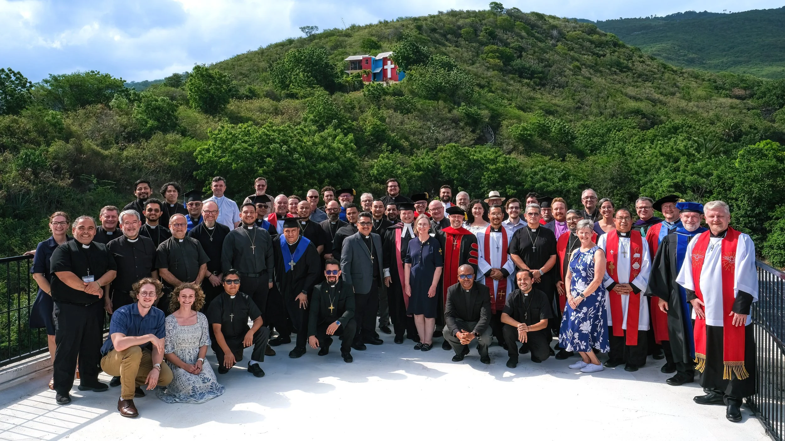 Grupo de personas, principalmente religiosas, posando en un balcón con paisaje montañoso y vegetación abundante, con una estructura de colores y una cruz en la cima de la colina en el fondo.