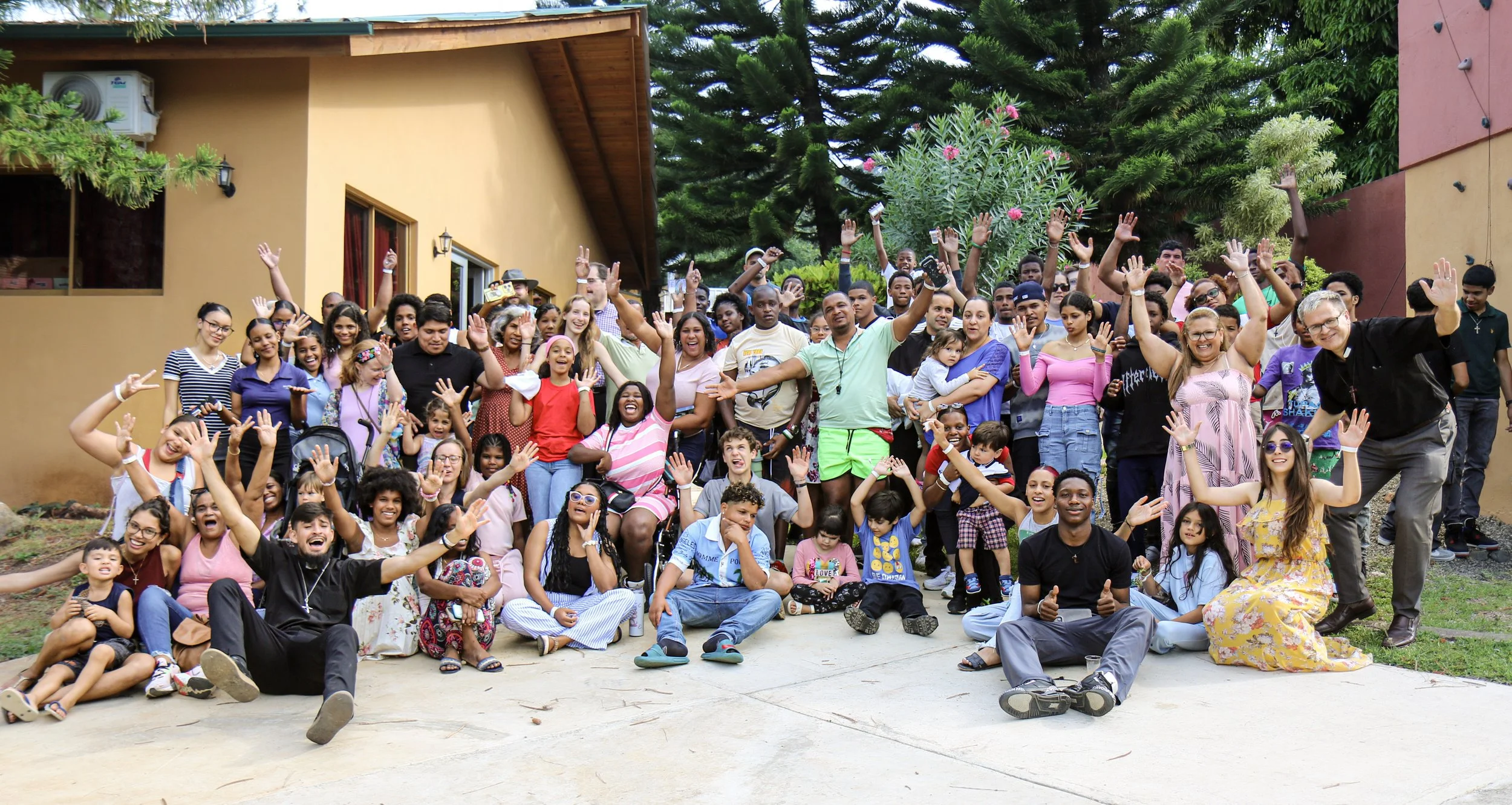 Un grupo diverso de personas, incluyendo niños y adultos, posan y sonríen en un área exterior con vegetación y árboles, celebrando con las manos levantadas.