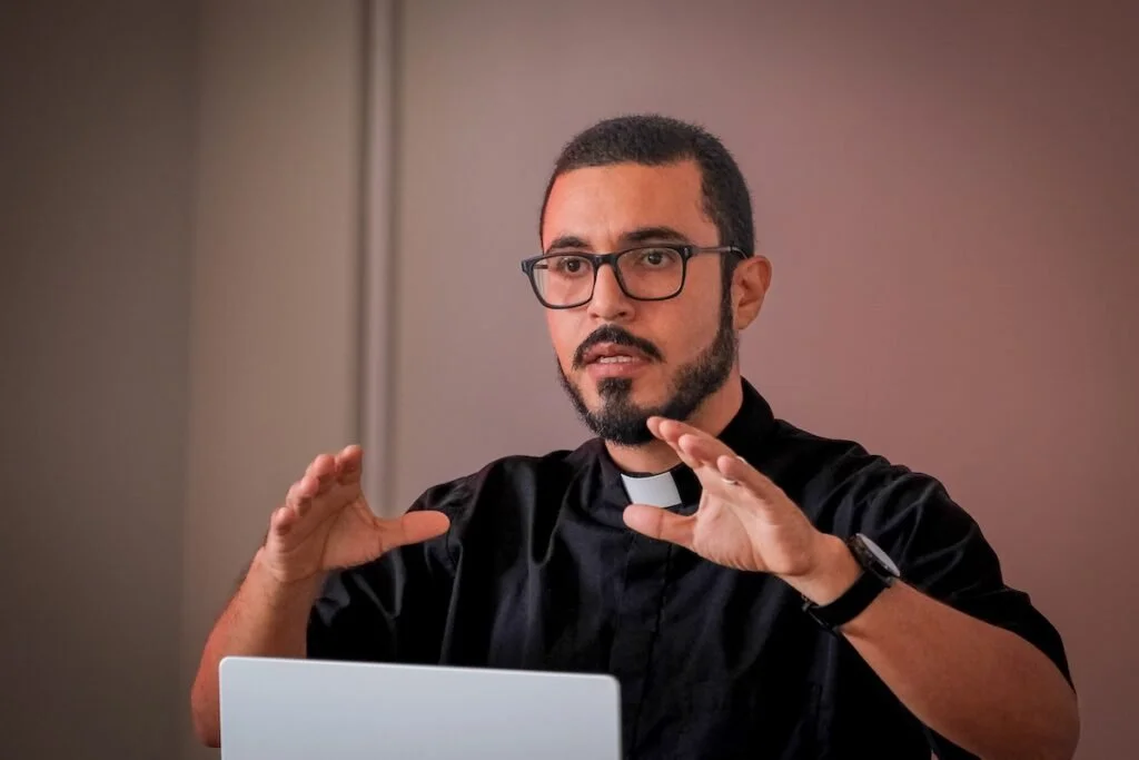 Un sacerdote con gafas y barba, vestido con sotana negra, hablando y gesticulando con las manos frente a una computadora.