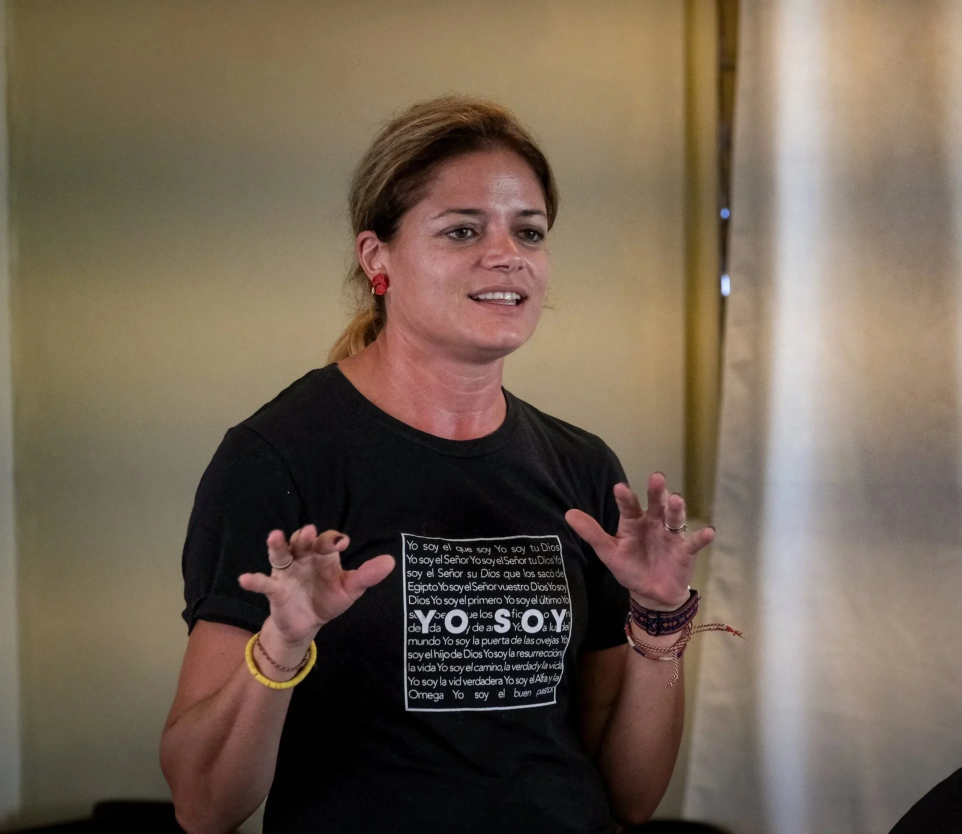 Mujer hablando con manos abiertas, lleva aretes rojos, pulseras y camiseta con texto religioso en español.