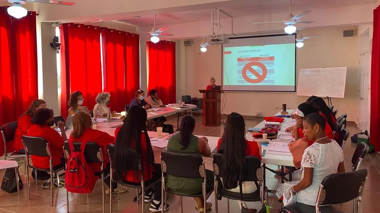 Una reunión o clase en un salón con varias personas sentadas en mesas en círculo, varias usam mascarillas, una presentación en pantalla y una pizarra blanca a la derecha.