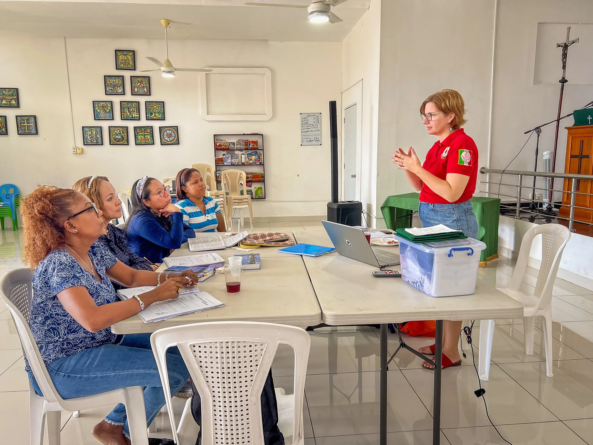 Mujer con gafas y camiseta azul con estampado, sentada en una silla blanca en una mesa, aparentemente participando en una reunión o clase, con varias otras personas también sentadas frente a ella. Una mujer de cabello corto, rubio y camisa roja está de pie frente a la mesa, hablando o presentando. El fondo muestra una pared con varias pinturas y un estante con libros.