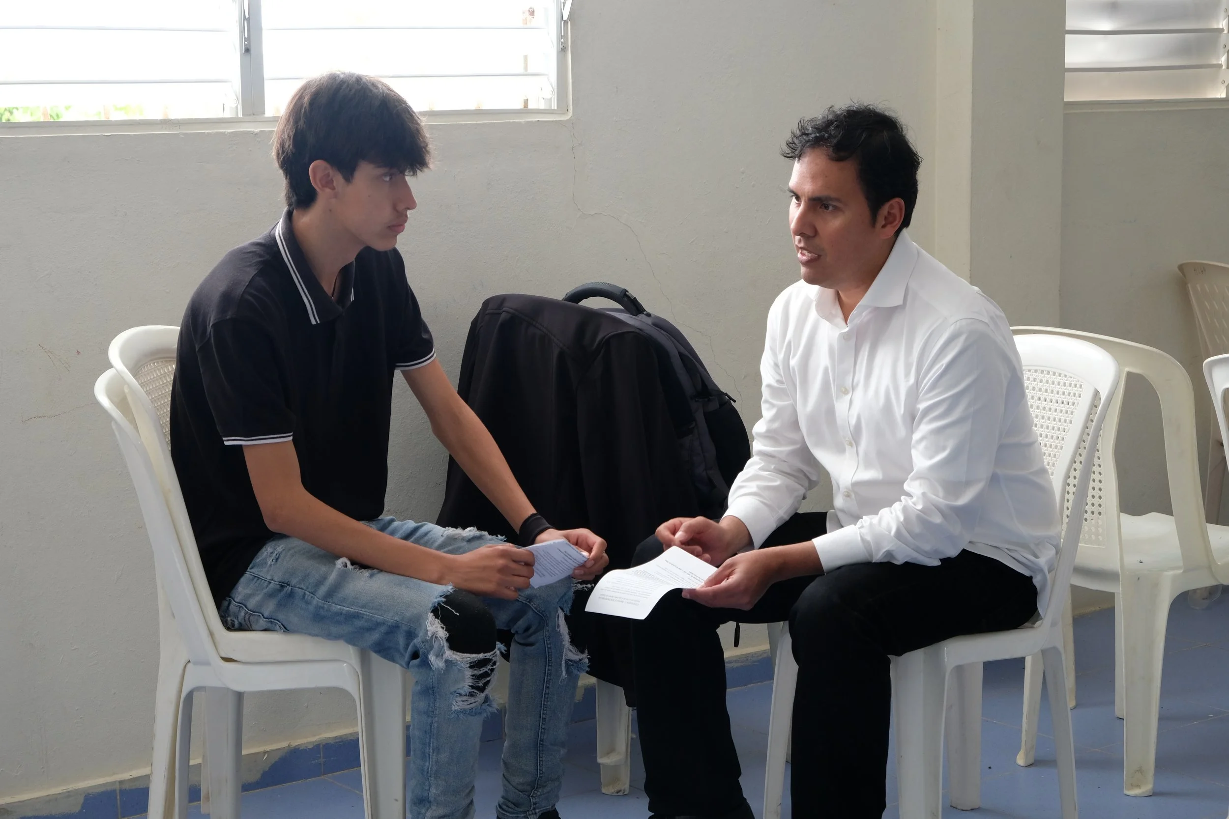 Un joven sentado en una silla blanca, vestido con camiseta negra con rayas en las mangas, sosteniendo un papel. Frente a él, un hombre vestido con camisa blanca, sentado en una silla similar, hablando con el joven. Ambos parecen tener una conversación seria, en un ambiente interior con paredes blancas y ventanas con persianas.