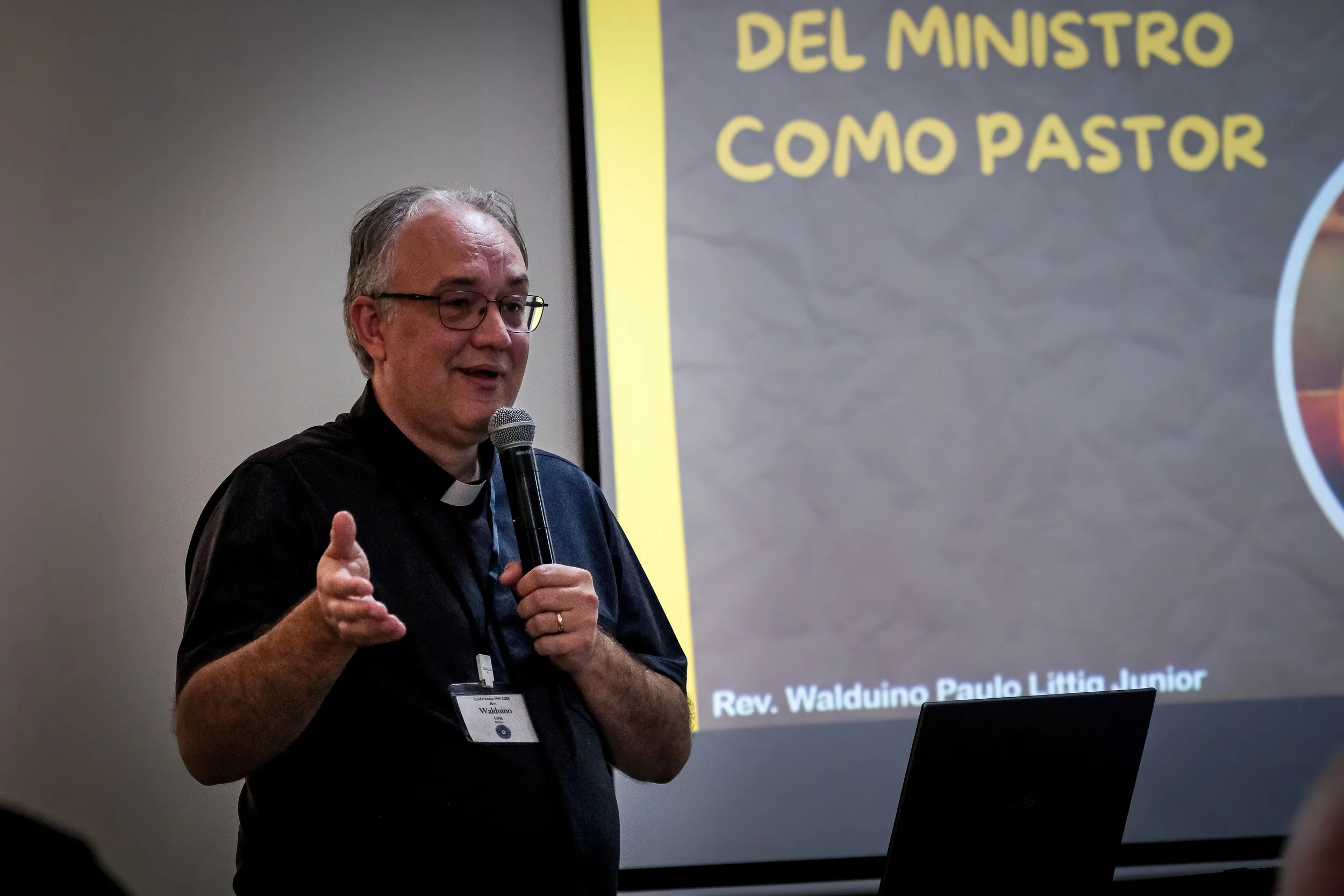 Hombre con gafas y camisa negra hablando en una conferencia, sosteniendo un micrófono, con una presentación en la pantalla detrás de él que dice 'Rev. Waldino Paulo Lúcio Junior' y en la parte superior 'DEL MINISTRO COMO PASTOR'.
