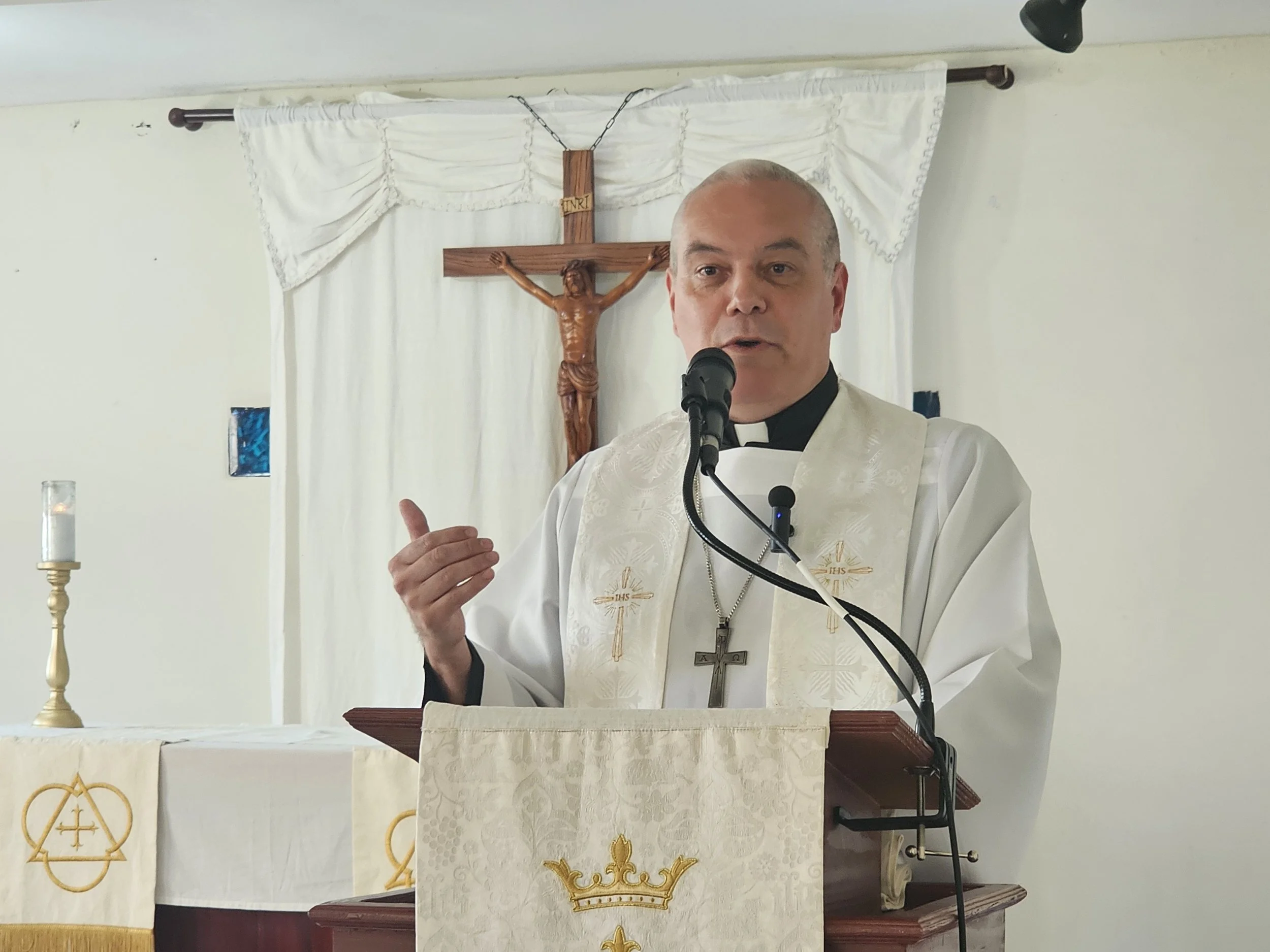Un sacerdote católico vestido con alba frente a un crucifijo en una iglesia, dando un sermón o una lectura, con una vela encendida a su izquierda.