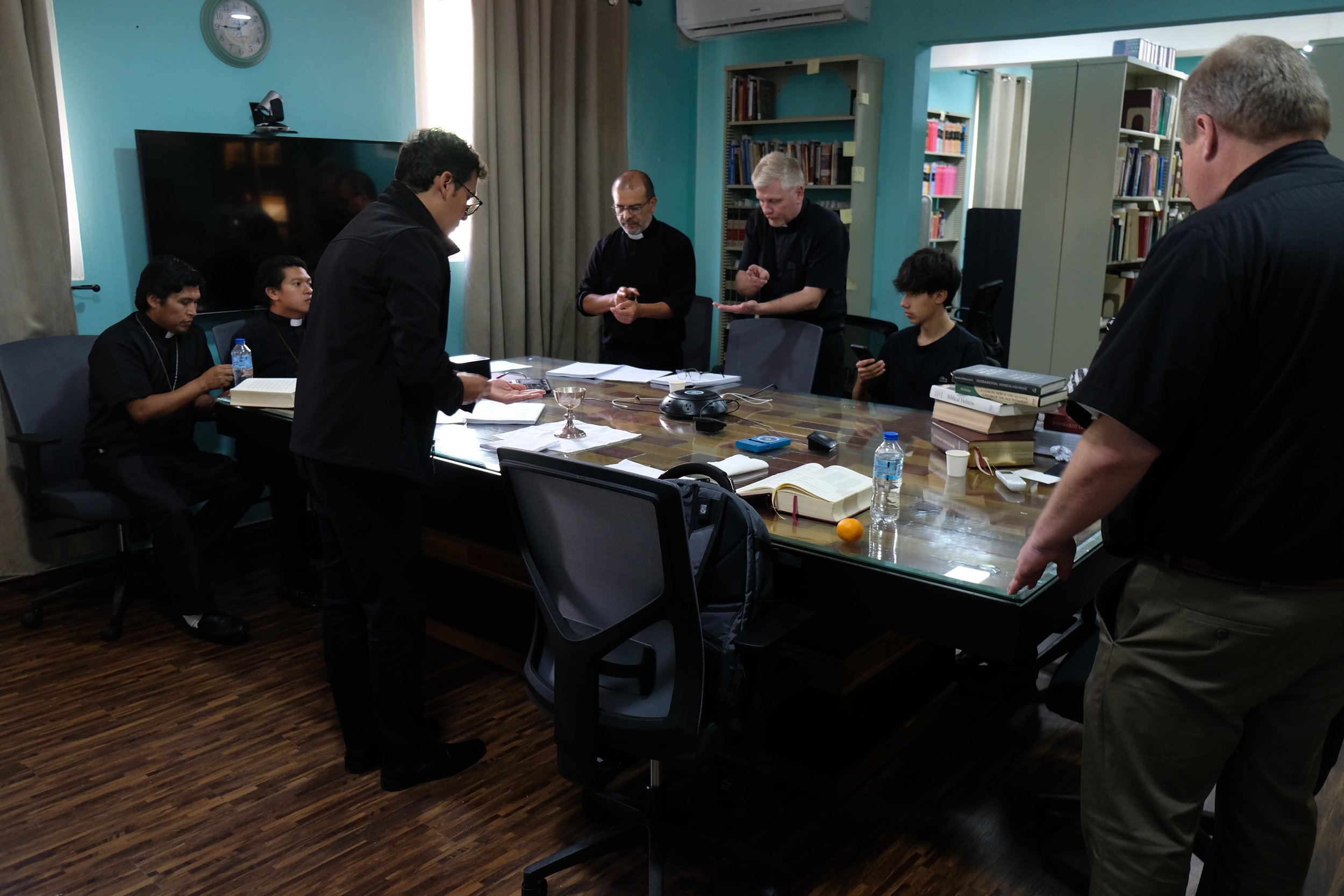Grupo de siete personas, algunas con hábitos religiosos, en una reunión en una sala de biblioteca con estantes llenos de libros, frente a una mesa de madera con objetos diversos, entre ellos un libro abierto y agua, en un ambiente formal.