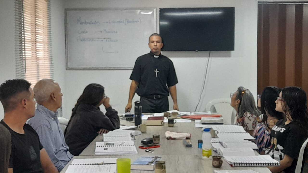 Un pastor luterano frente a una pizarra y un televisor en una sala de reuniones, rodeado de varias personas sentadas en una mesa de conferencias con libros, cuadernos y tazas.