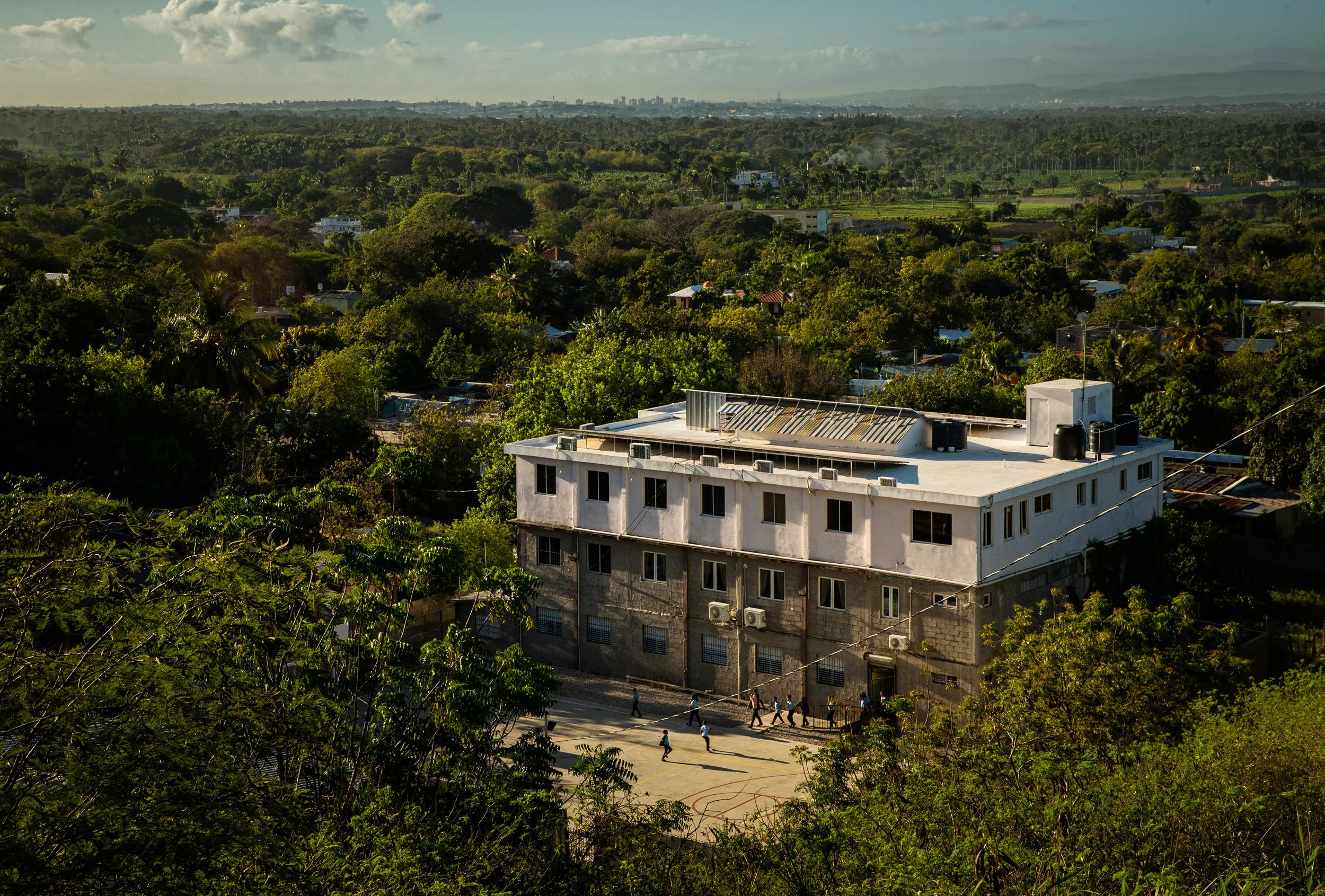 Vista de una ciudad rodeada de áreas verdes con árboles, casas, y edificios en el horizonte bajo un cielo con nubes.