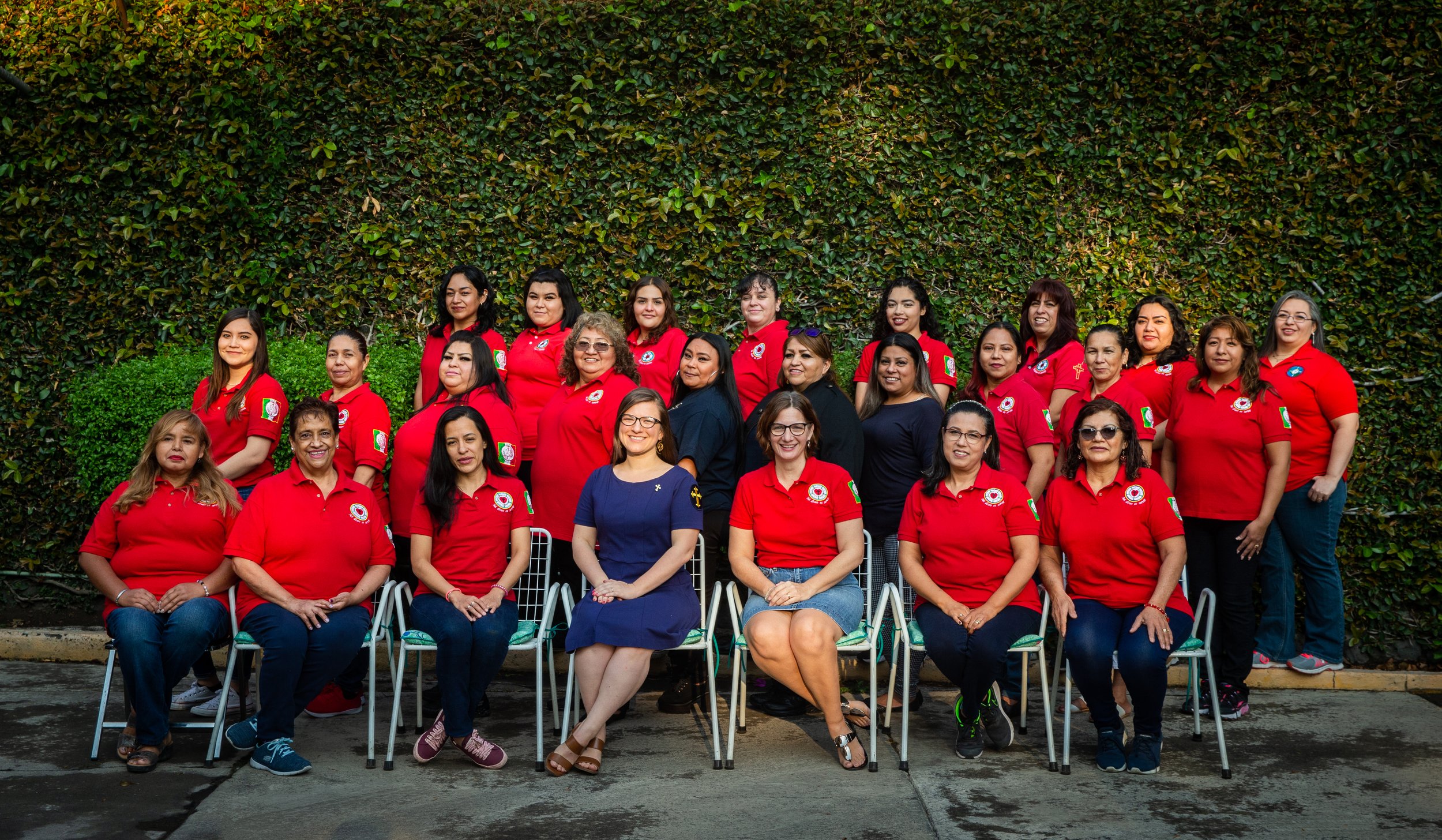 Grupo de mujeres y unas cuantas en el frente, todas con camisetas rojas, posando para una foto grupal frente a un muro de arbustos verdes.