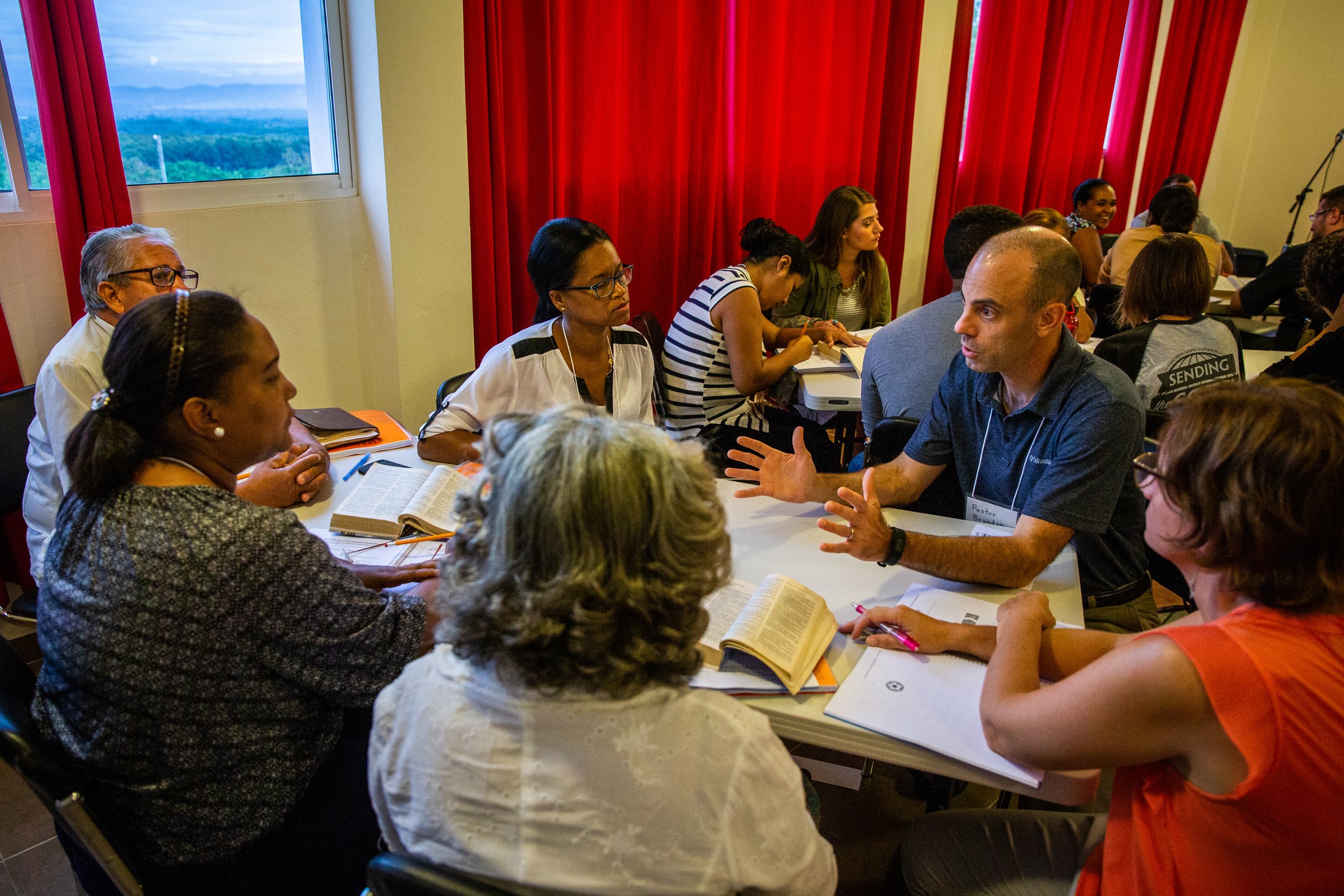 Un grupo de personas participando en una reunión o taller en una sala con cortinas rojas y una ventana que muestra un paisaje de montañas y cielo azul.