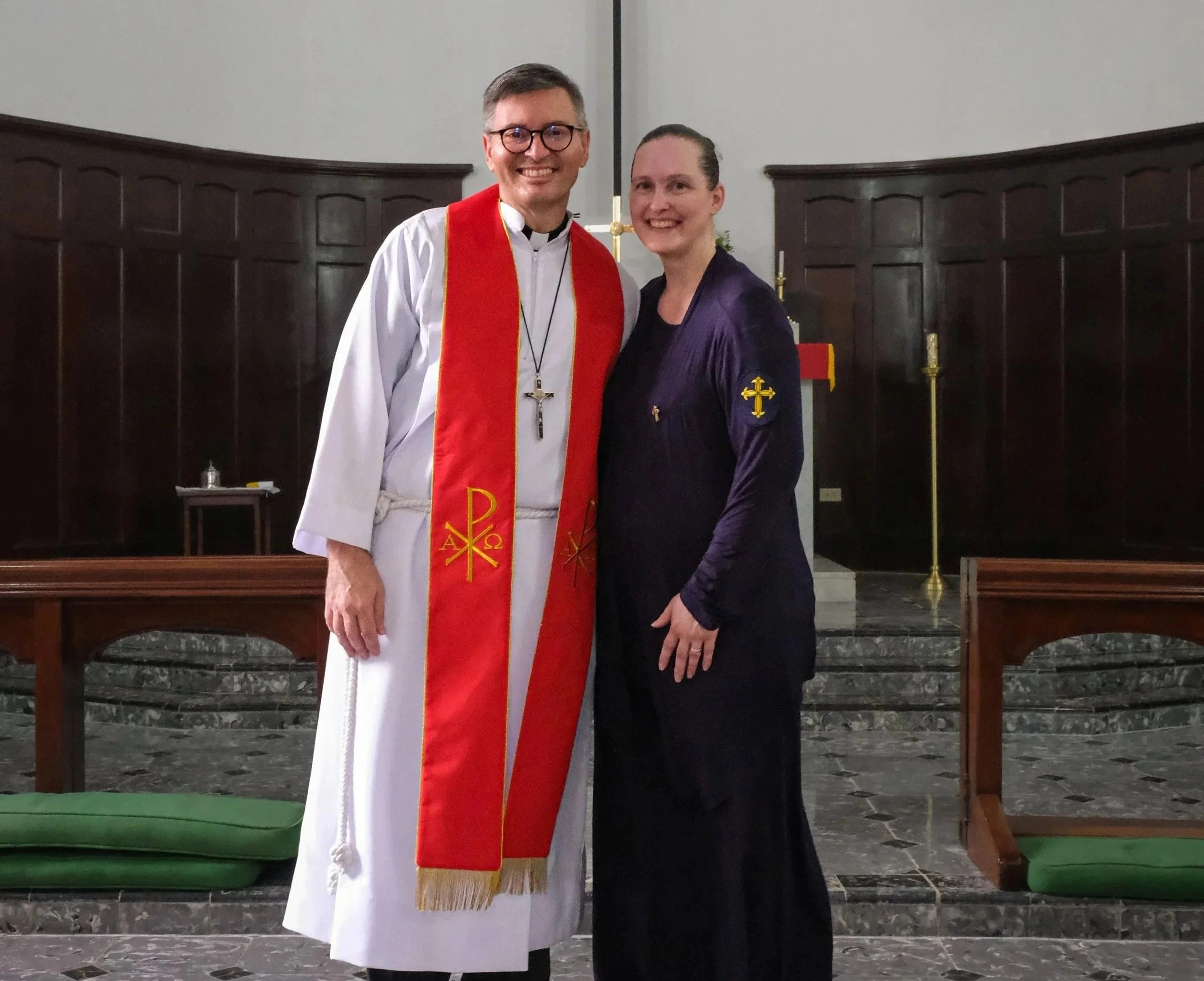 Dos personas en una iglesia durante una ceremonia religiosa. Uno vestido con toga blanca y estola roja con símbolos religiosos, la otra con vestido negro y blazer. Ambos son sonriendo y de pie frente a una cruz y velas.