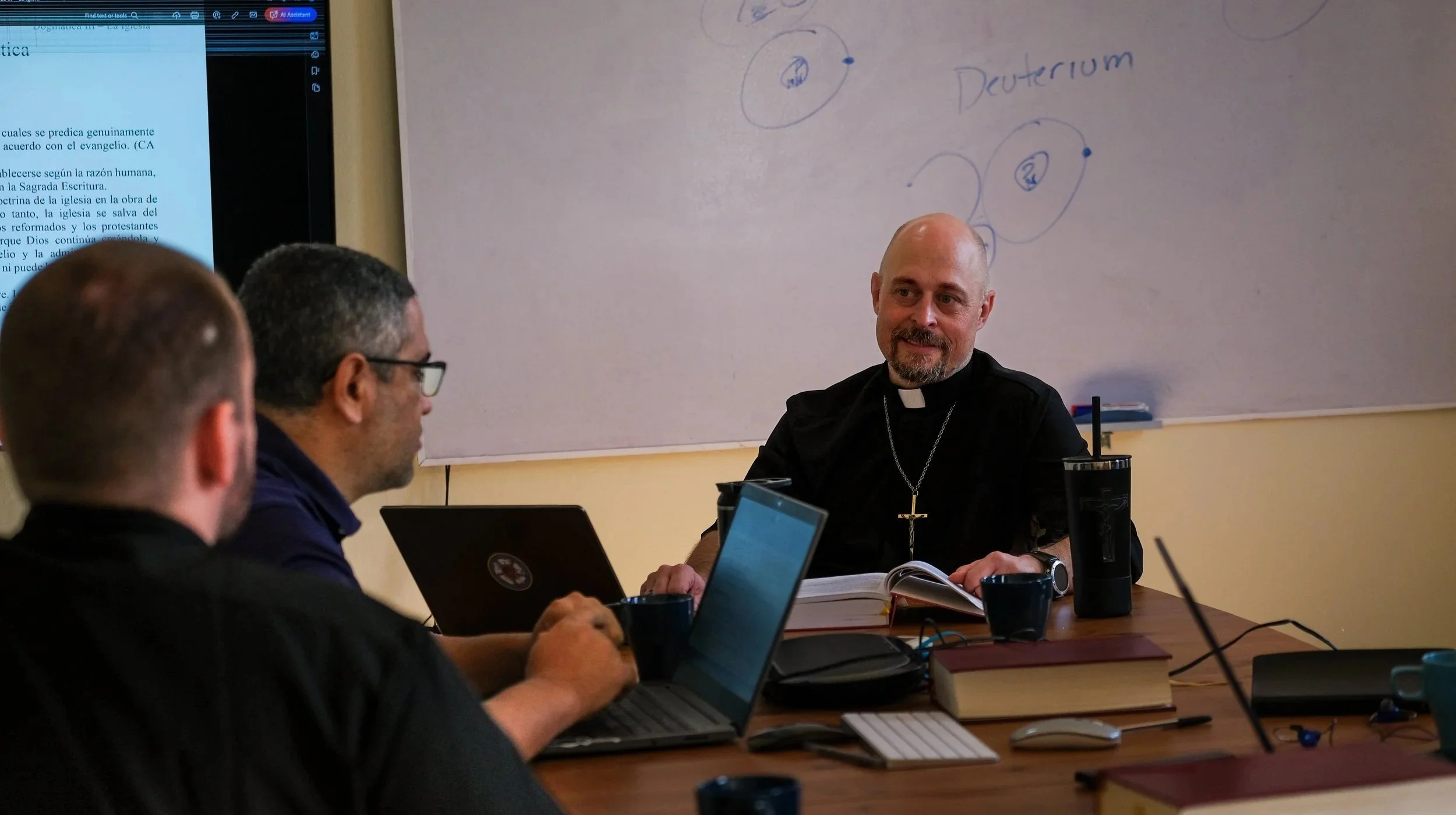 Un grupo de personas en una sala de reuniones, incluyendo a un sacerdote con crucifijo, que está hablando. En la pared, hay una pizarra con notas y diagramas. Dos personas usan laptops, y hay varios libros y vasos en la mesa.