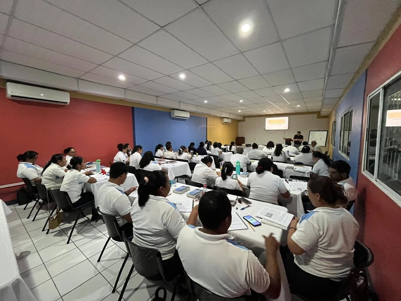 Grupo de personas asistiendo a un curso o conferencia en un aula con pantallas y pizarras.