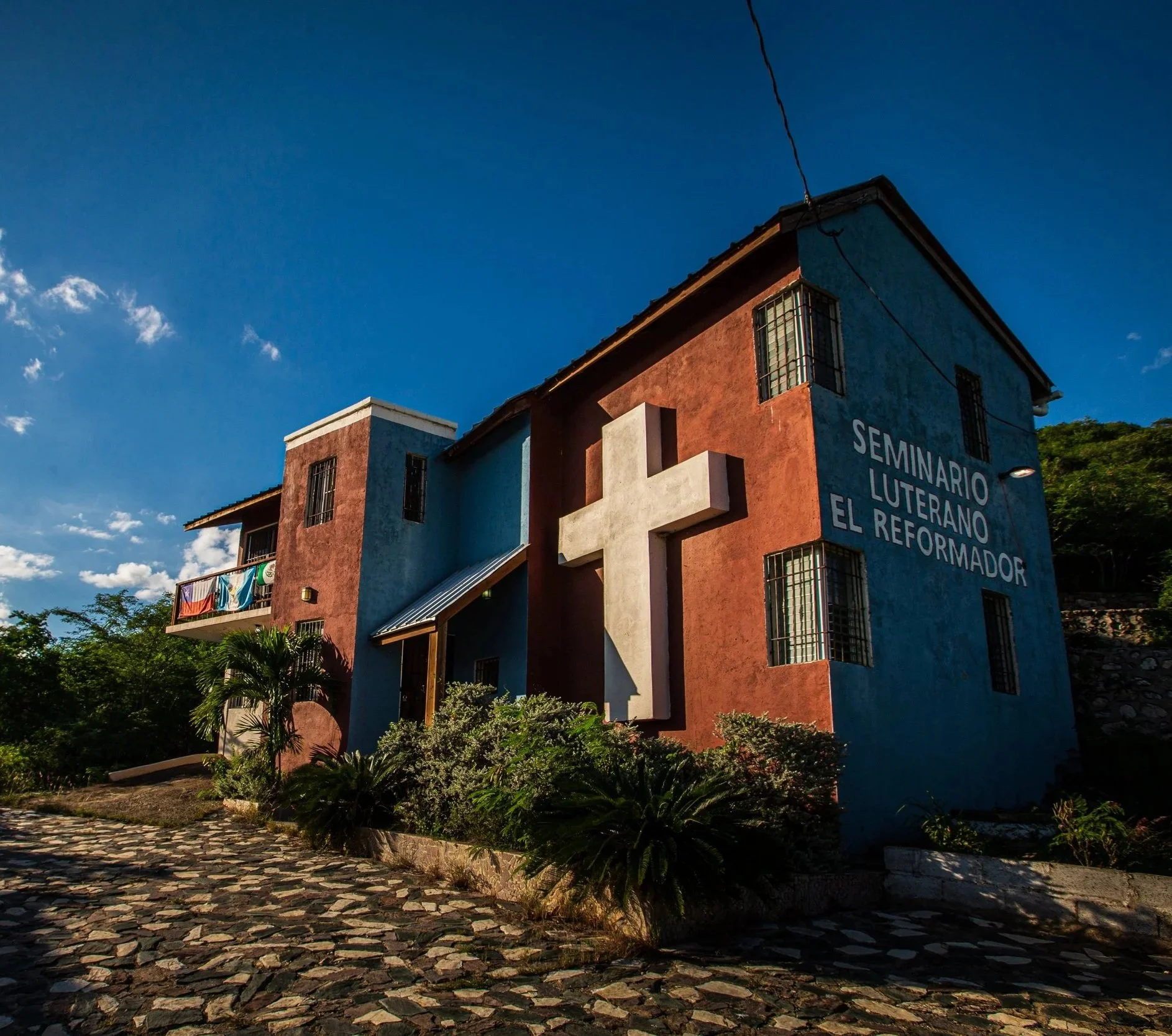 Edificio colorido con signos que indican un seminario religioso, rodeado de plantas y un camino empedrado, bajo un cielo despejado.