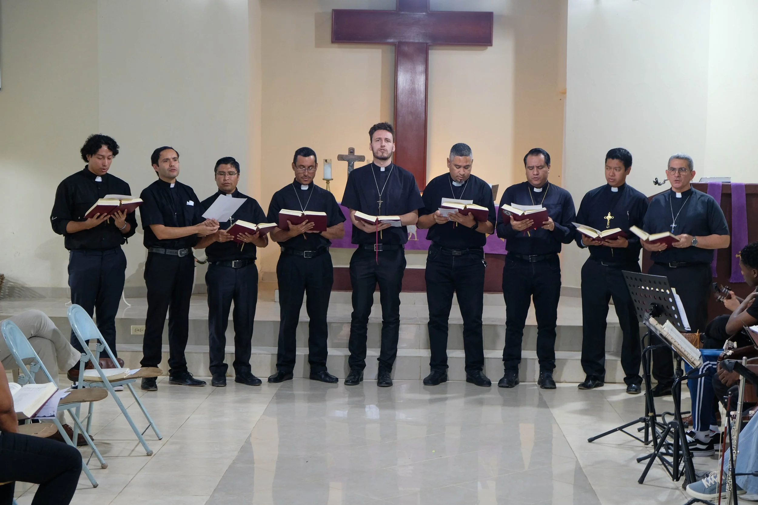 Un grupo de hombres en túnicas negras, algunos con cruces, están de pie en un altar en una iglesia, participando en un canto o oración.
