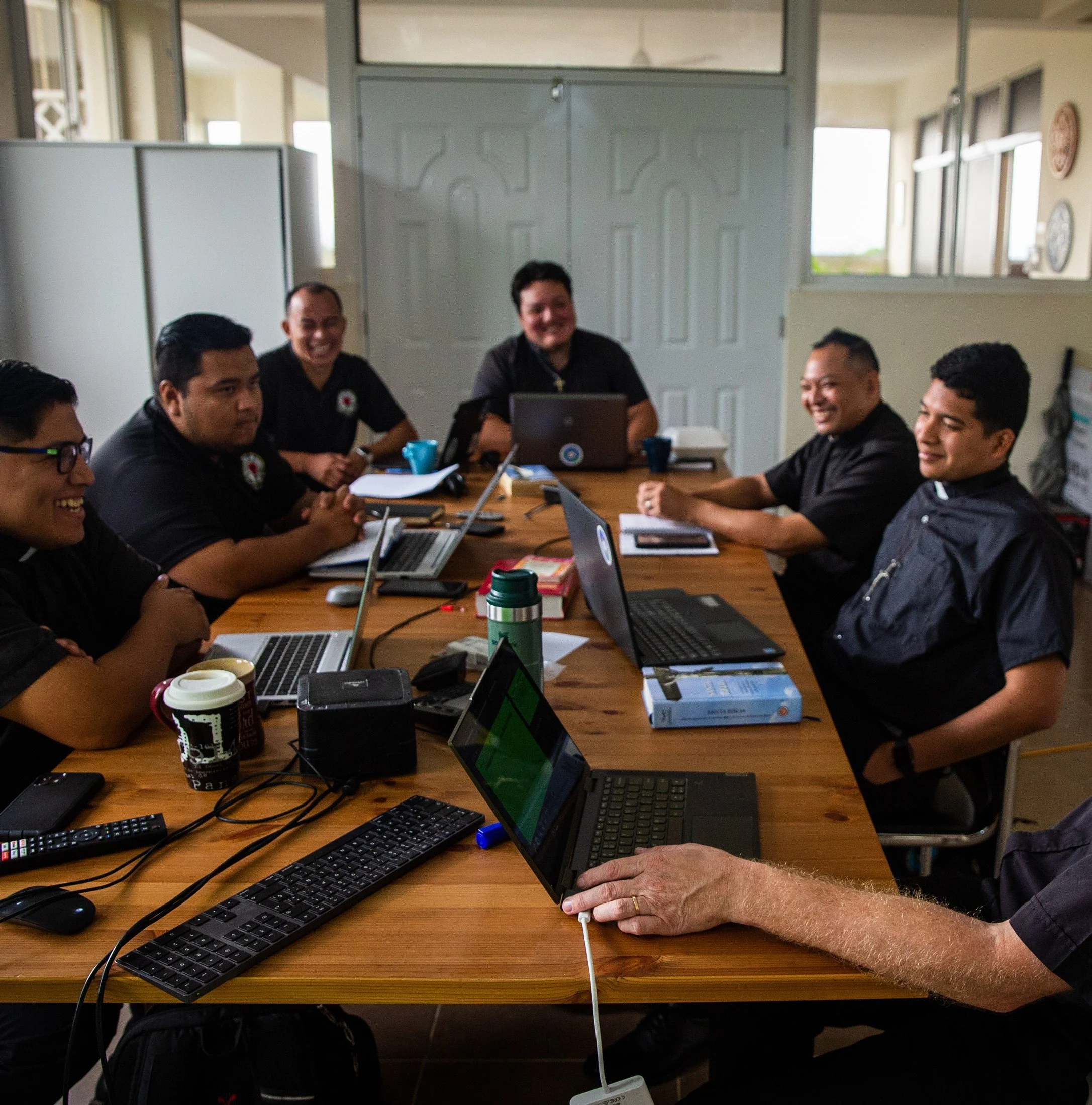 Reunión de varias personas en una sala de oficina, la mayoría viste uniforme negro y algunos usan laptops y papeles sobre la mesa.