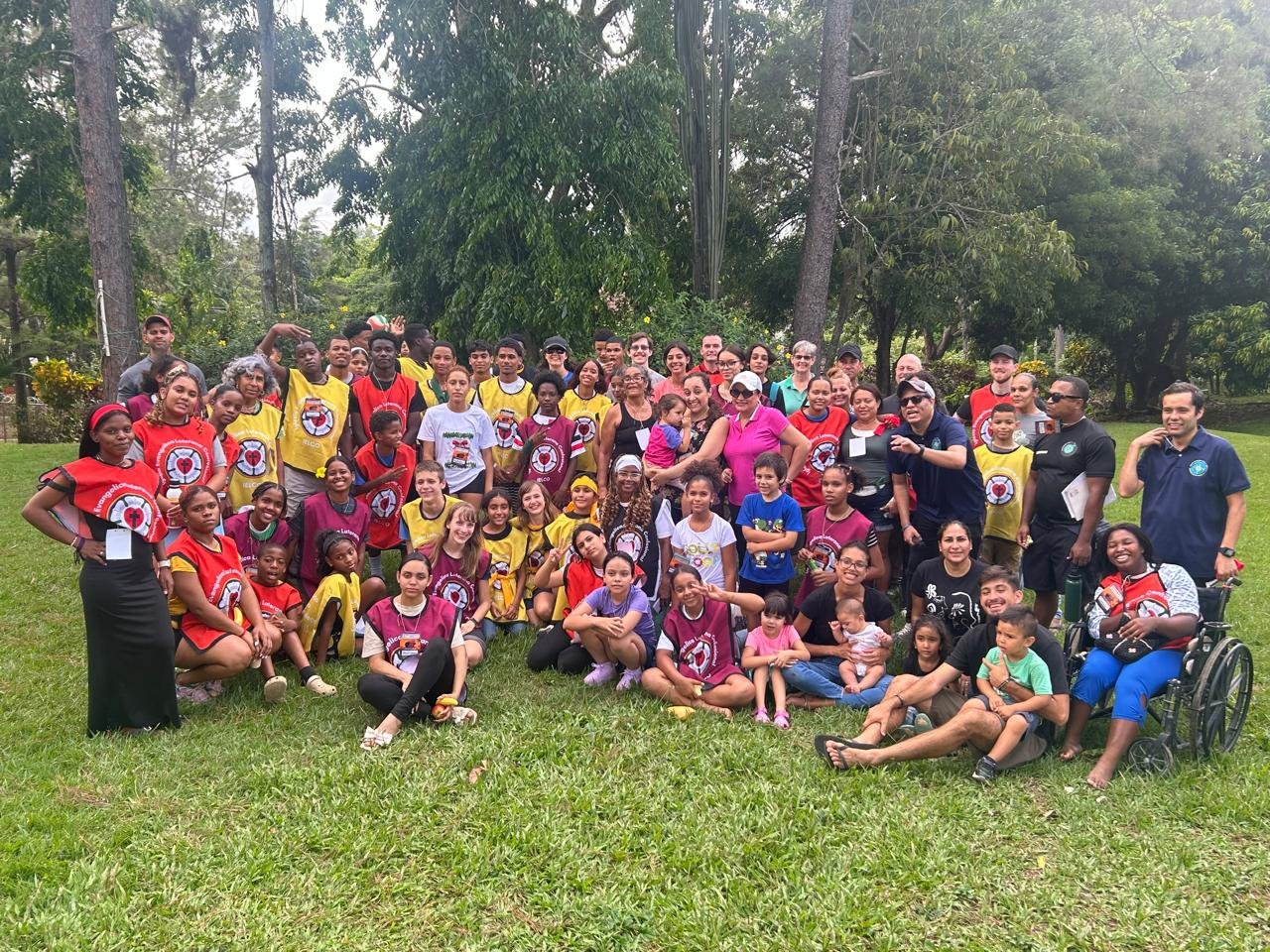 Grupo grande de personas, niños y adultos, en un parque, algunos con camisetas de diferentes colores y un fondo de árboles.