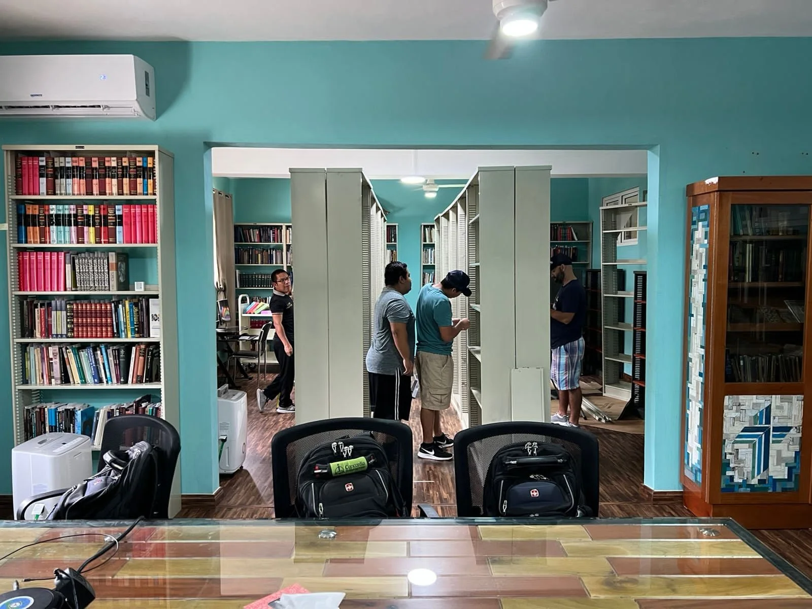 Varias personas dentro de una biblioteca, revisando libros en estanterías. Hay sillas con mochilas en primer plano y un aire acondicionado en la pared. Ambiente tranquilo y organizado.