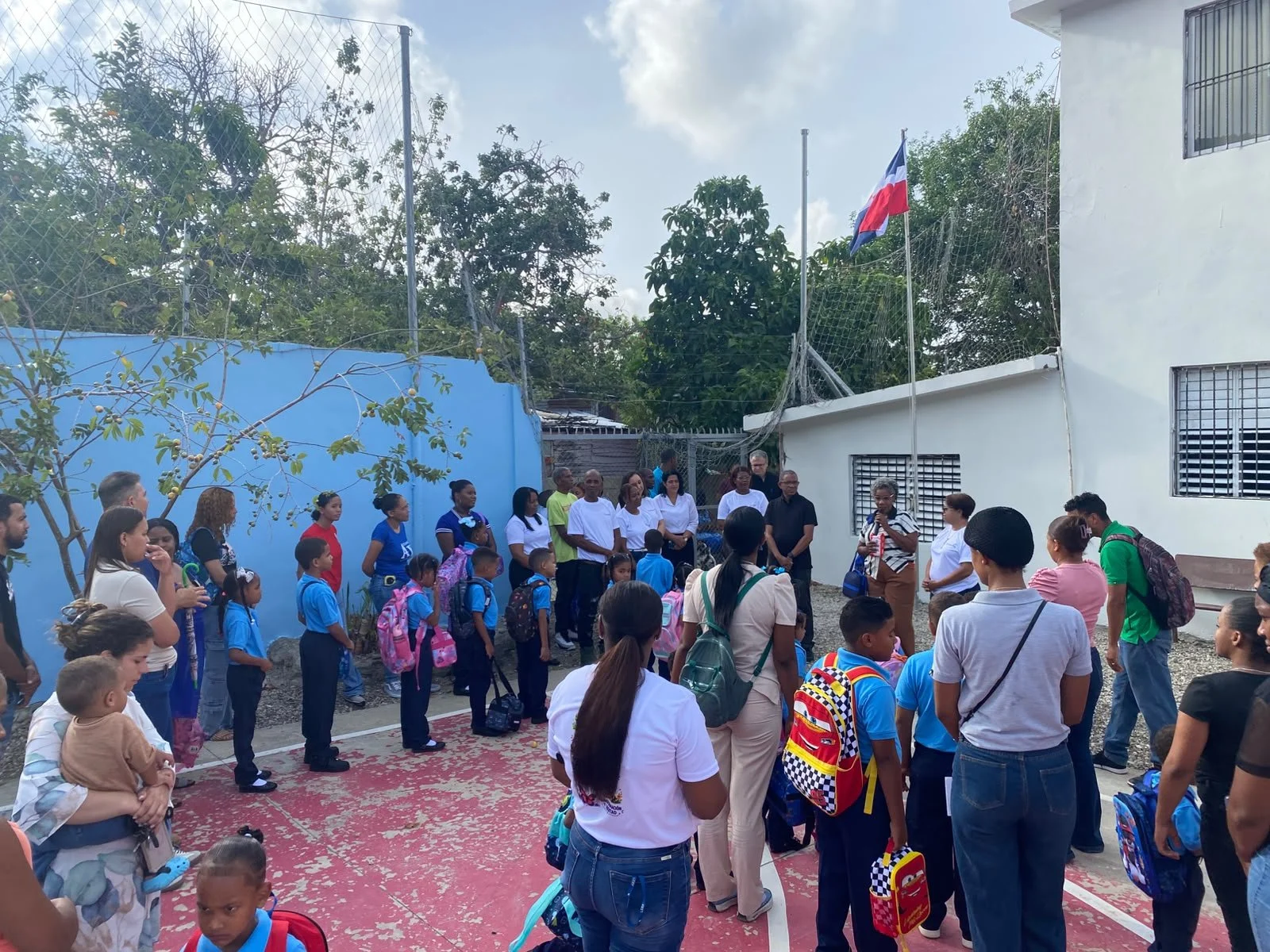 Grupo de niños y adultos en una escuela, algunos con mochilas, en un ambiente exterior con paredes blancas y azul, una cancha de deporte y banderas en el fondo.
