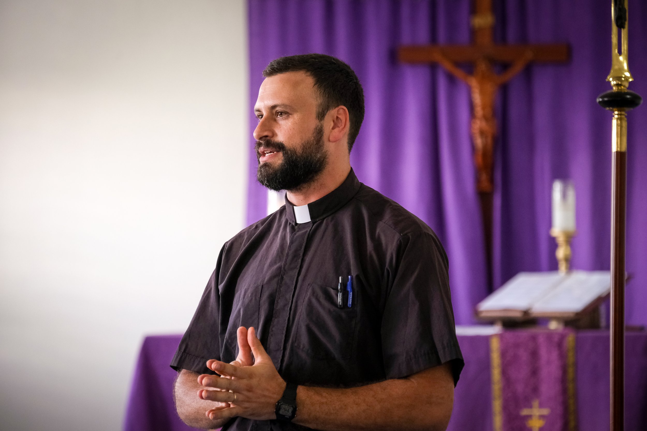 Un sacerdote en una iglesia, con hábitos clericales negros y un collarín blanco, de espaldas en oración, frente a un fondo de cortinas moradas con una cruz de madera y candelabros dorados.