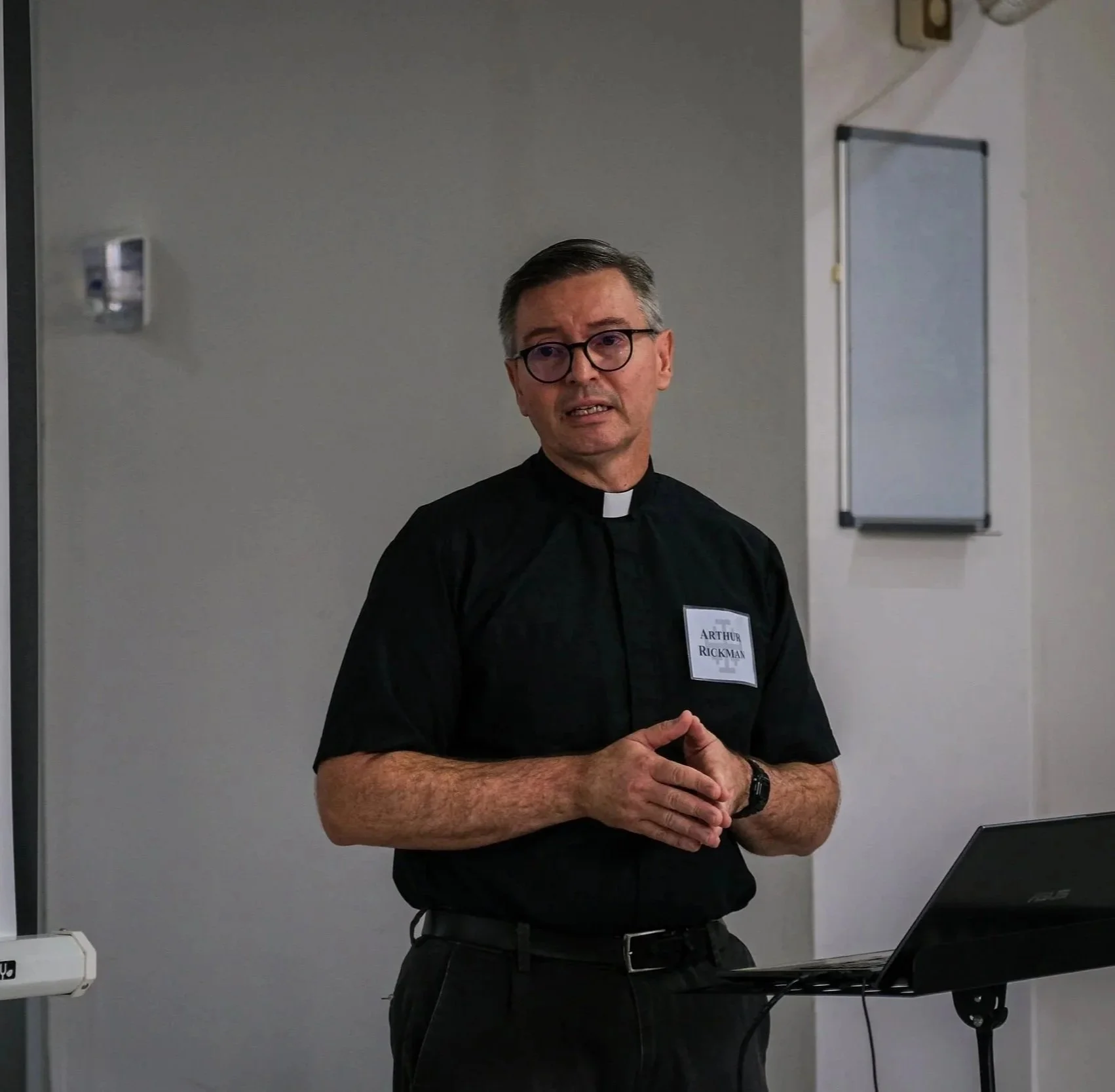 Hombre vestido con collar clerical, gafas, y camiseta negra, hablando en una sala de clases o reunión. Tiene un nombre pegado en su ropa que dice 'Arthur Rickman'.