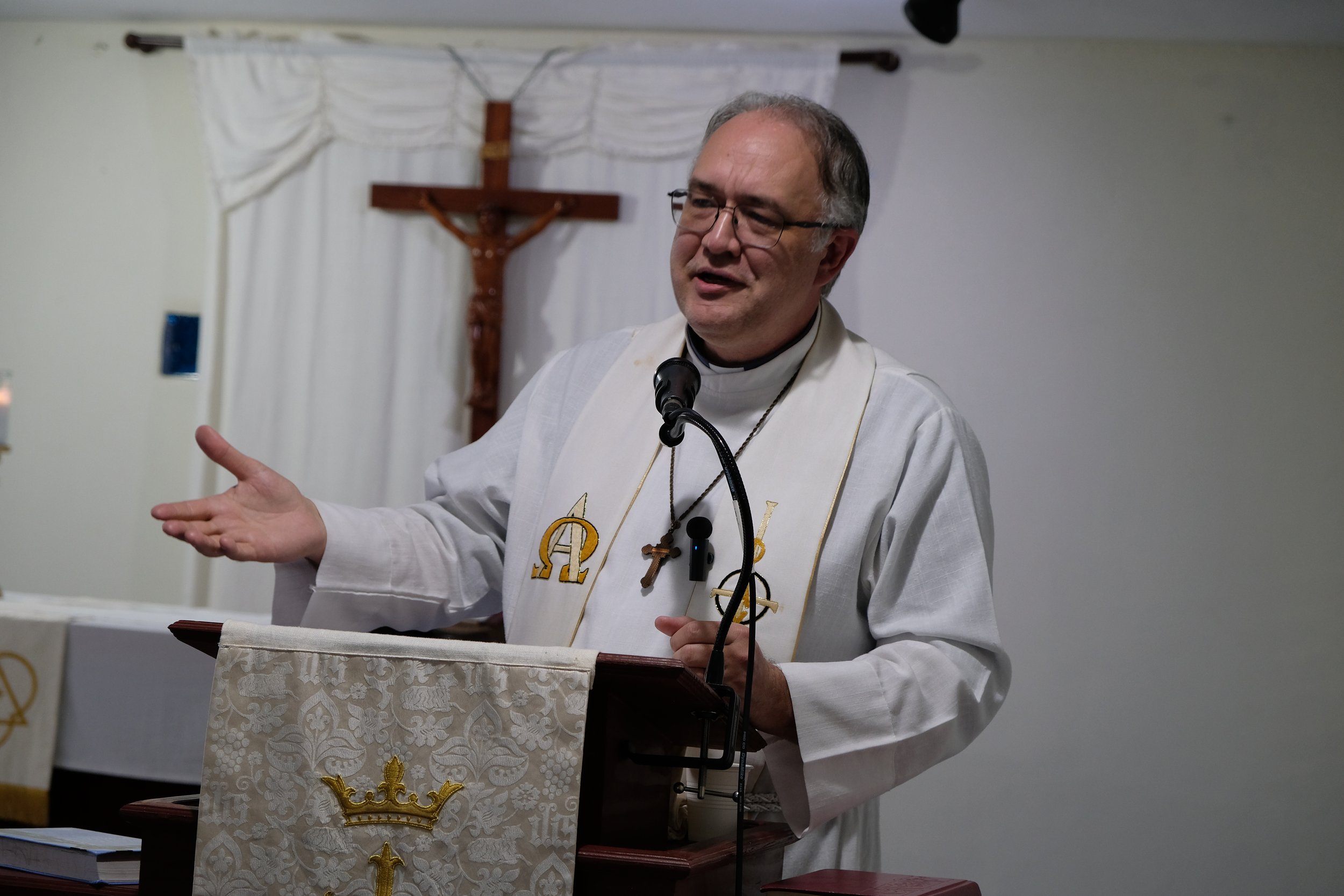 Un sacerdote vestido con vestimenta blanca, con símbolos religiosos en el pecho, dando un sermón o discurso en una iglesia delante de un atril decorado, con una cruz en la pared y cortinas blancas en el fondo.