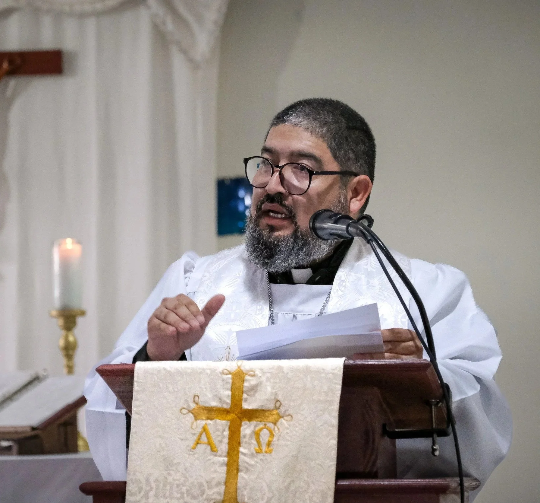 Un pastor luterano con barba y gafas, vestido con atuendo religioso, hablando en un micrófono y sosteniendo unos papeles, en un entorno de iglesia con cortinas blancas y una vela en el fondo.