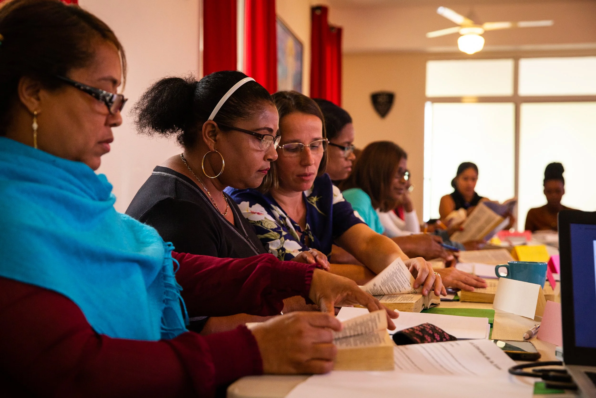 Grupo de mujeres sentadas en una mesa, leyendo libros y revisando documentos, en un ambiente interior con ventanas grandes y cortinas rojas.