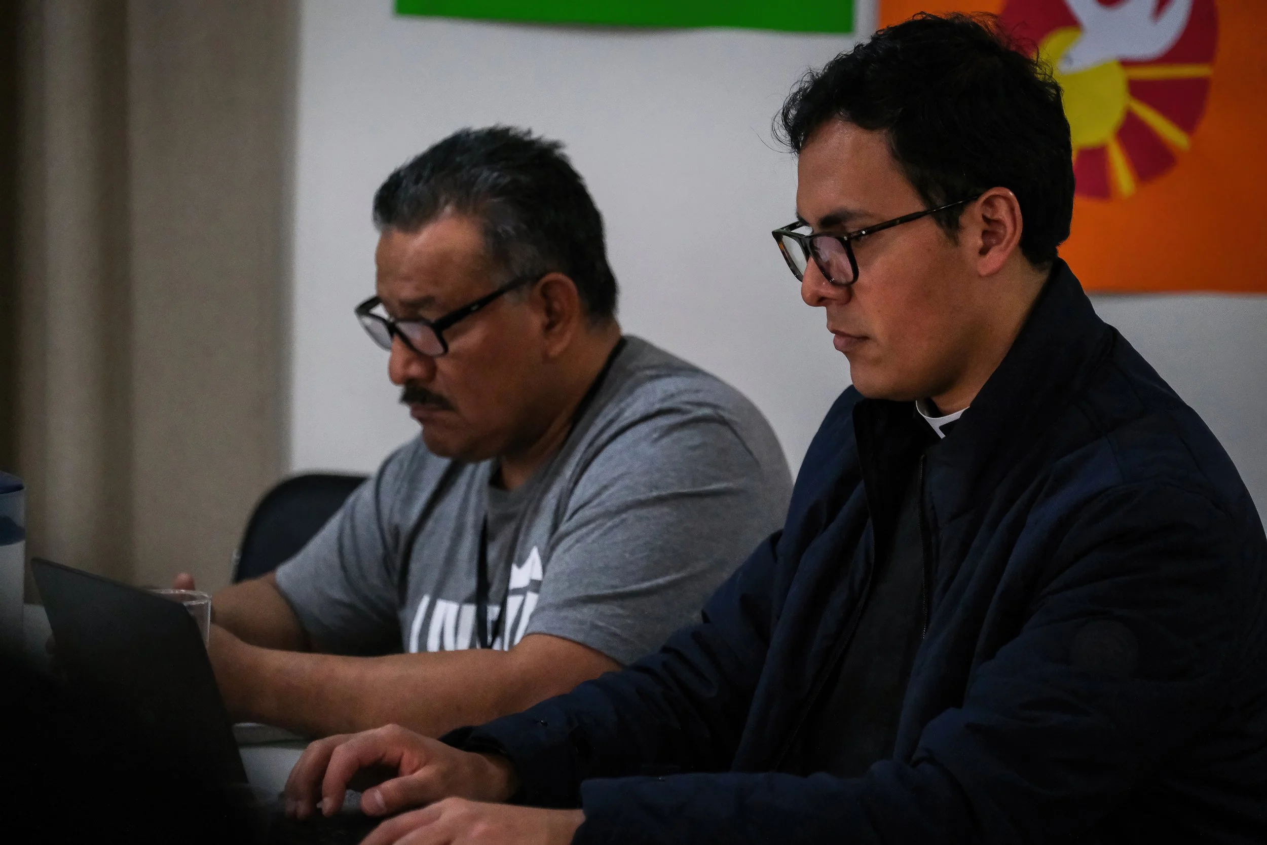 Dos estudiantes con gafas miran hacia abajo mientras están sentados en una mesa, uno usando una chaqueta azul oscuro y el otro una camiseta gris con rayas blancas y negras, en un entorno interior.