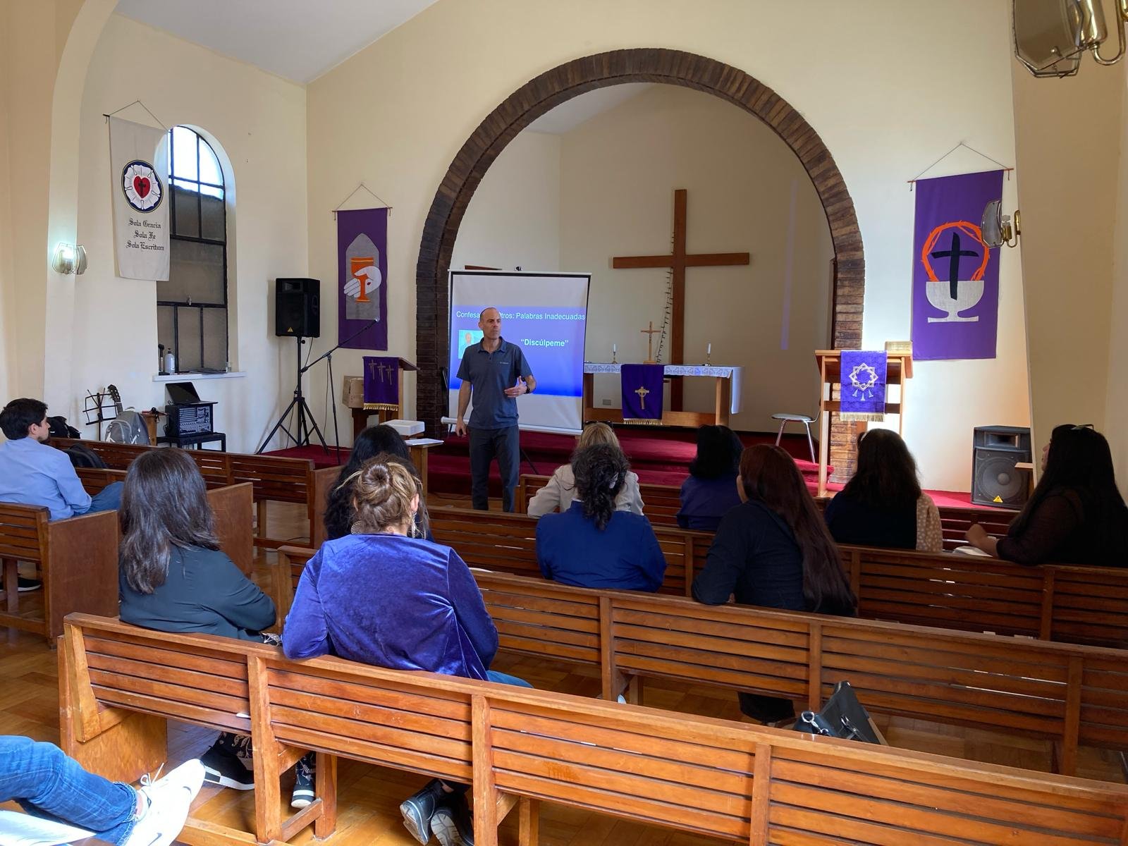 Vista del interior de una iglesia con personas sentadas en bancas escuchando a un hombre que da una charla frente a una pantalla proyectada y atrás un altar con una cruz. Hay banderas religiosas en las paredes.