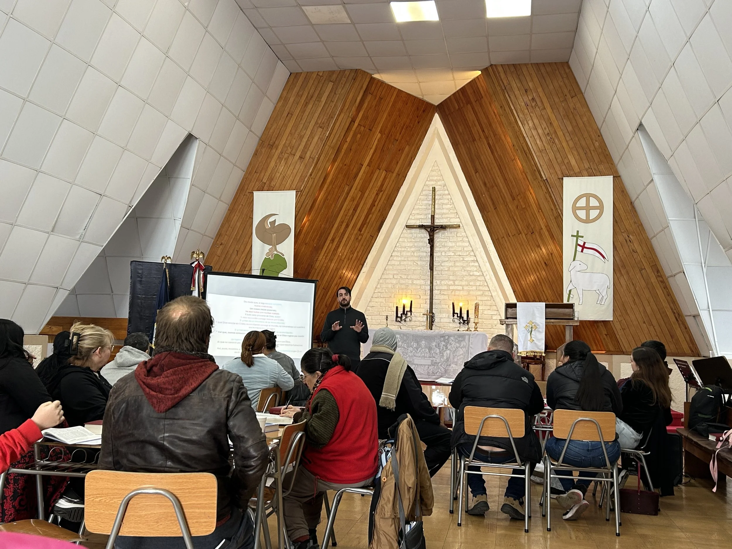 Una iglesia con altar de madera y pared de ladrillos, con personas sentadas en sillas de madera en una reunión o misa.
