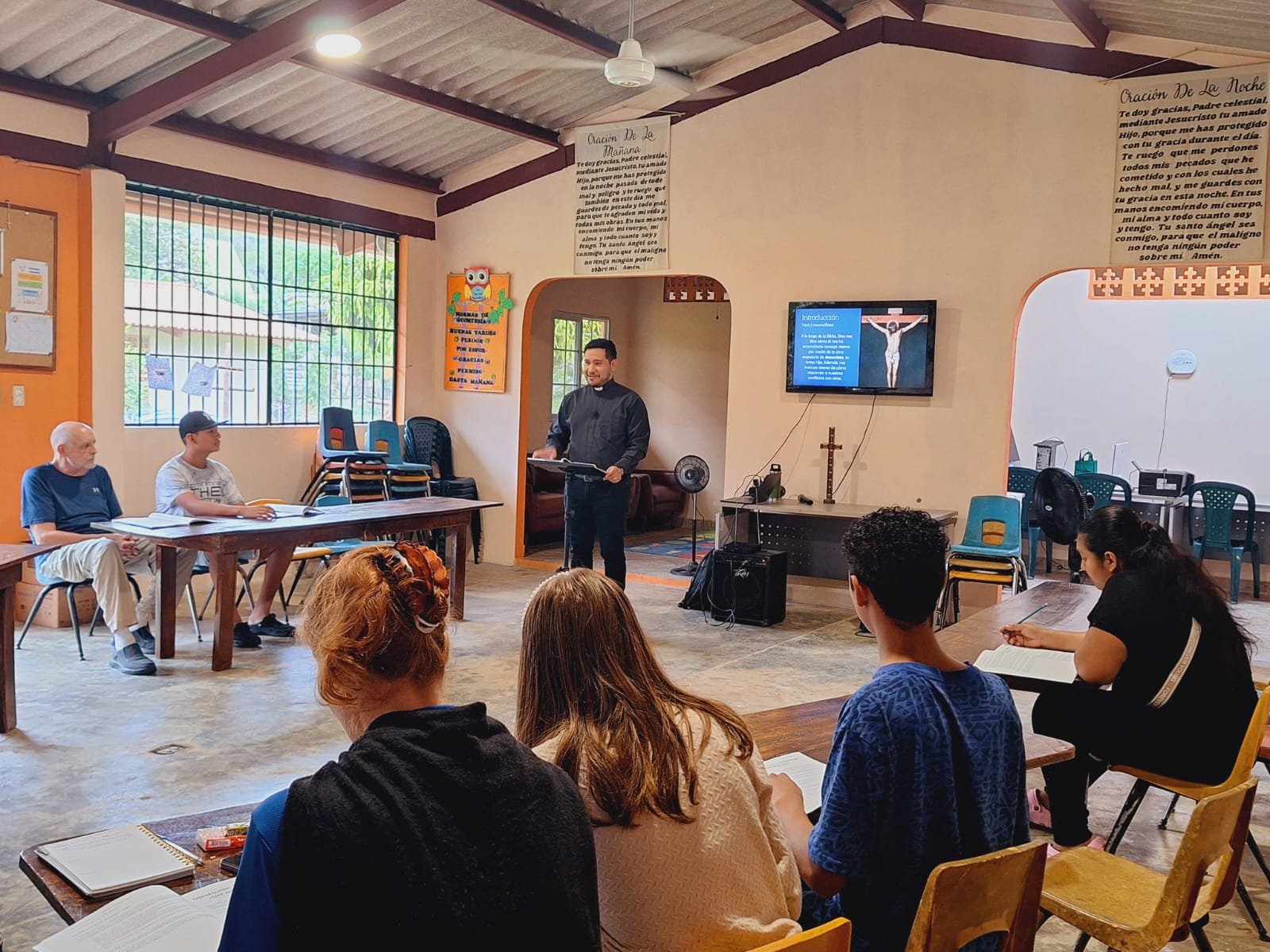 Reunión religiosa en un salón con personas sentadas, un presentador de pie, pantalla con imagen de Jesús crucificado y pasos de un estudio bíblico.