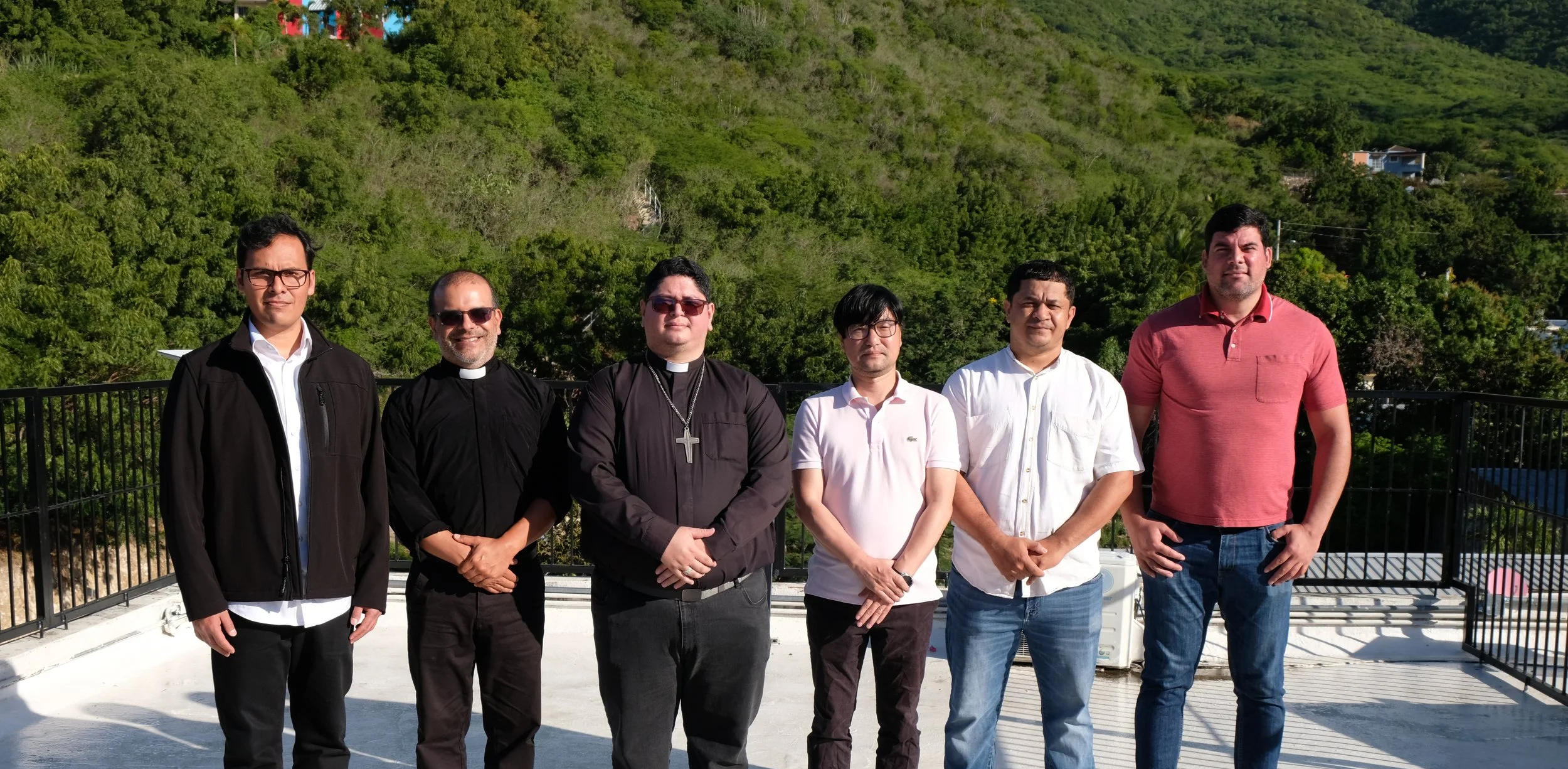 Foto de seis hombres frente a un fondo de colinas verdes y árboles, algunos con vestimenta formal y otros con ropa casual, en un lugar al aire libre con una baranda negra.