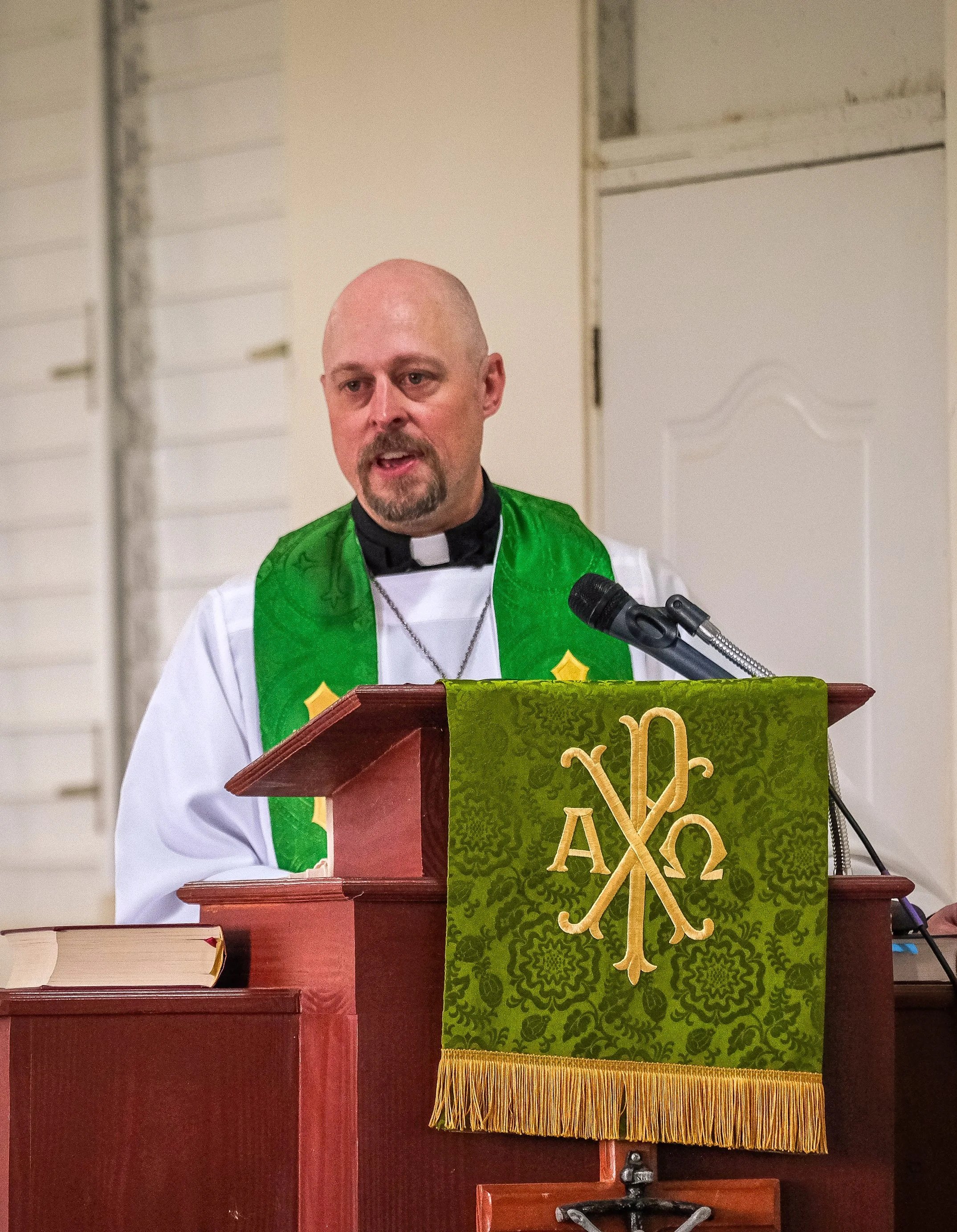 Un pastor luterano habla en un podio decorado con símbolos religiosos, en una habitación con paredes blancas y un libro sobre una mesa a su lado.