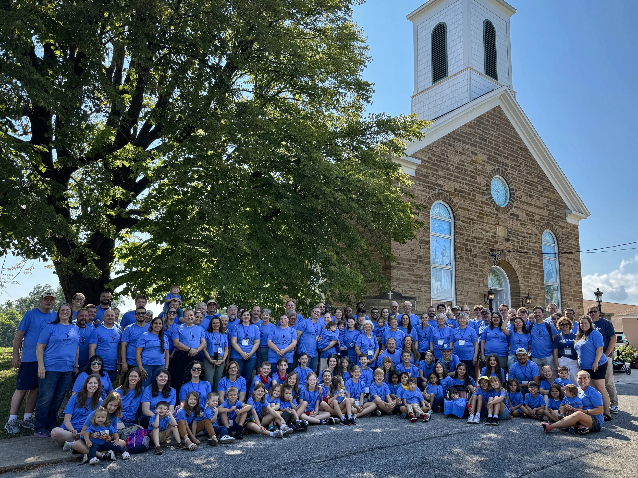 Un grupo grande de personas, en su mayoría niños y algunos adultos mayores, posando frente a una iglesia de ladrillo con ventanas de arco y un campanario, en un día soleado y con árboles verdes de fondo. Todos llevan camisetas azules.
