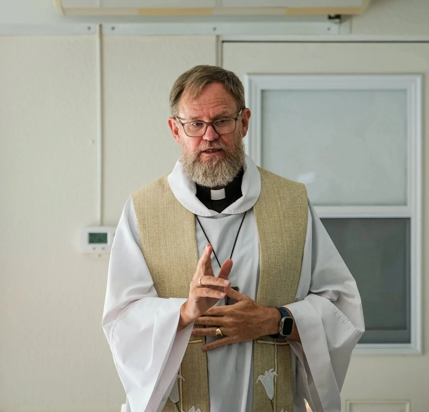 Un pastor luterano con barba y gafas, vistiendo sotana blanca y sudario beige, haciendo un gesto con los dedos, en un entorno interior con paredes blancas y vidrios.