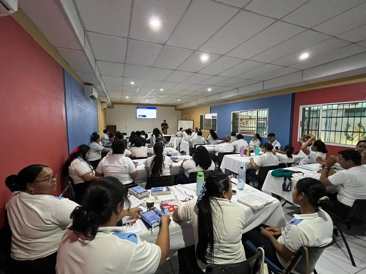 Sala de aula con estudiantes sentados en mesas cubiertas con manteles blancos, escuchando a un profesor en el frente y tomando notas.