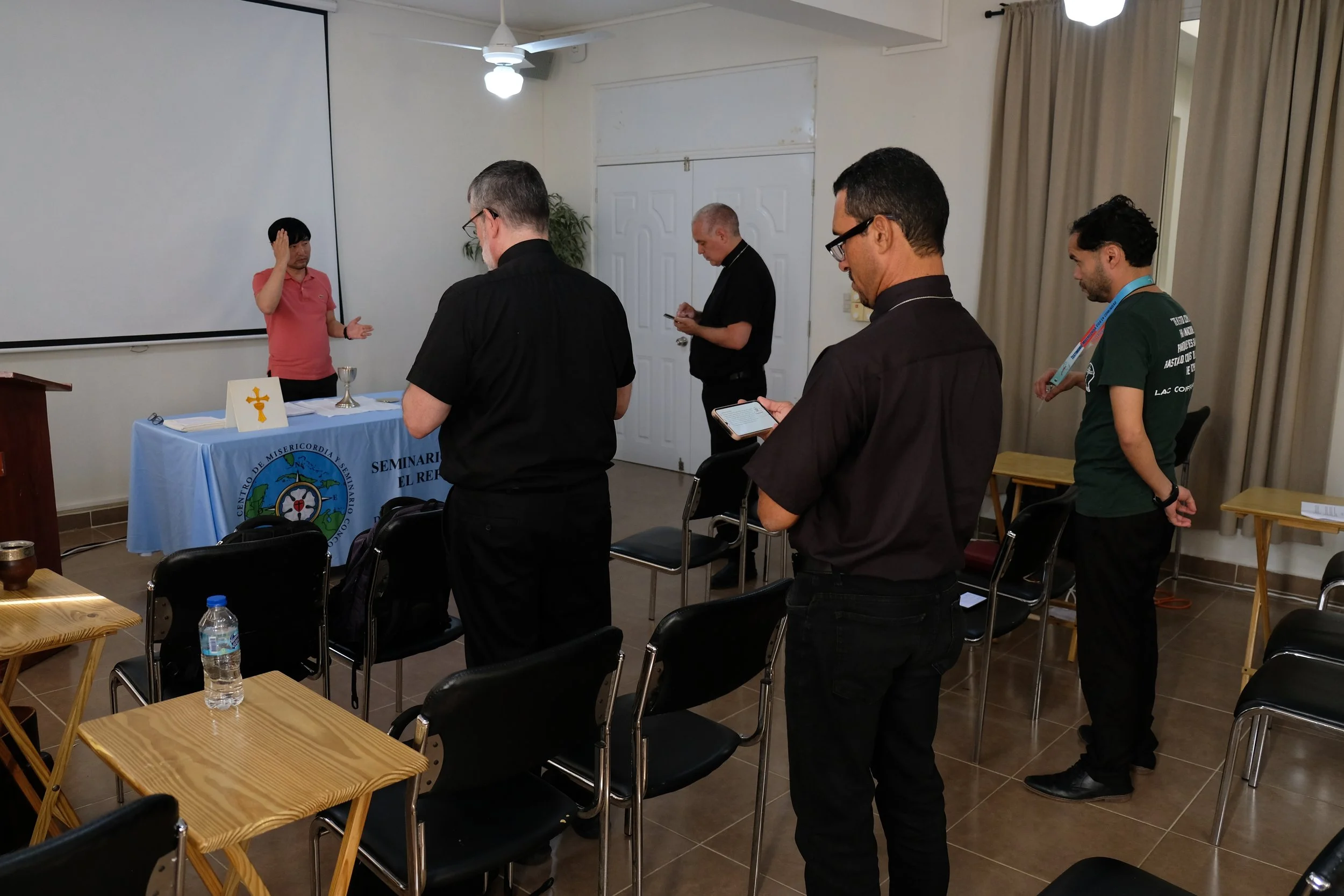Un grupo de personas participa en una ceremonia religiosa, con una persona levantando la mano en señal de bendición y una mesa con símbolos religiosos y una copa, en un salón con cortinas y sillas.
